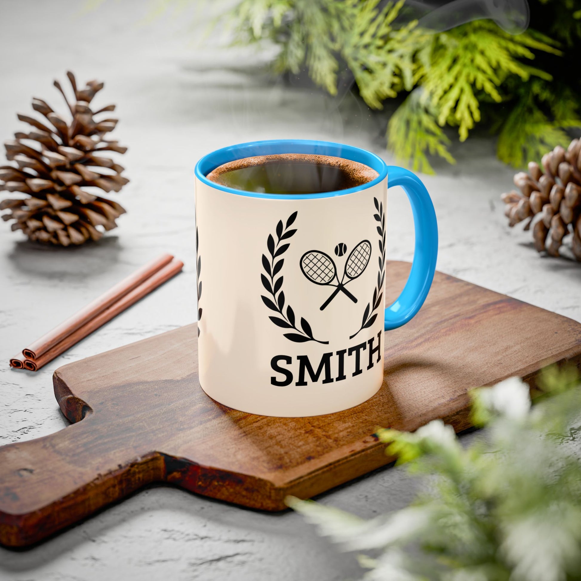 Tennis Coffee Mug A ceramic coffee mug with a cream-colored body, bright blue handle and rim, and a black tennis racket graphic with the name 'SMITH' printed below. The mug sits on a wooden cutting board surrounded by pinecones, cinnamon sticks, and green foliage on a gray surface.