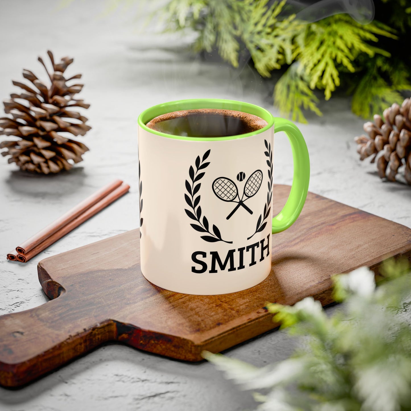 Tennis Coffee Mug A ceramic mug with a cream-colored exterior, a bright green handle and interior rim, and a black tennis racket graphic with the name 'SMITH' printed below. The mug sits on a wooden cutting board surrounded by pinecones, cinnamon sticks, and green foliage on a gray surface.