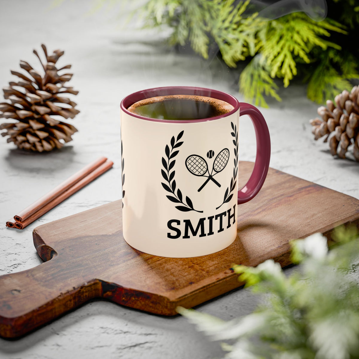 Tennis Coffee Mug A cream-colored ceramic mug with a dark red handle and rim, featuring a black graphic of crossed tennis rackets under a laurel wreath and the name 'SMITH' printed below. The mug is filled with a hot beverage and sits on a wooden cutting board surrounded by pinecones, cinnamon sticks, and green foliage on a gray stone surface.