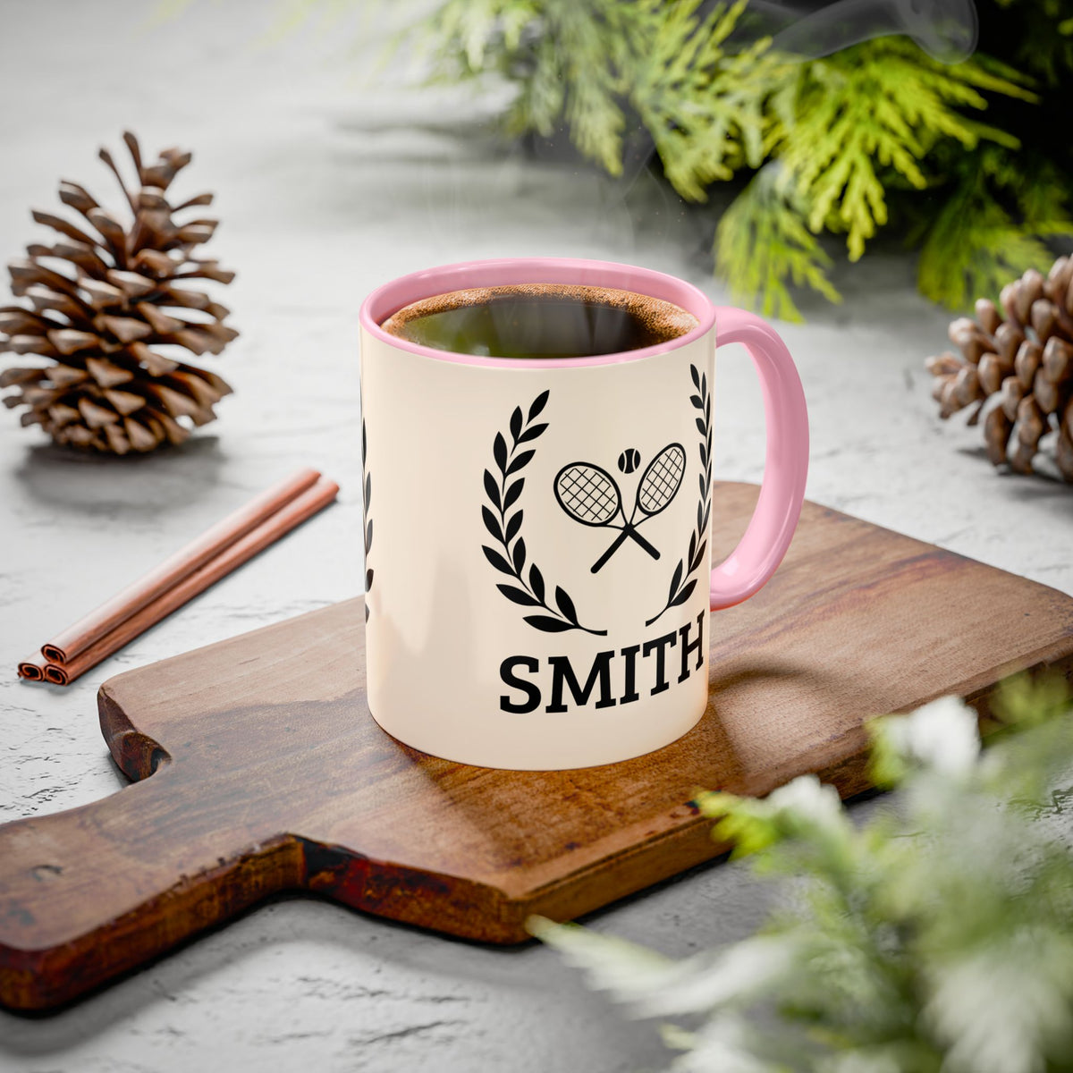 Tennis Coffee Mug A personalized ceramic coffee mug with a pink handle and interior, featuring a black tennis racket and laurel wreath graphic above the name 'SMITH'. The mug sits on a wooden cutting board surrounded by pinecones, cinnamon sticks, and green foliage on a gray stone surface.
