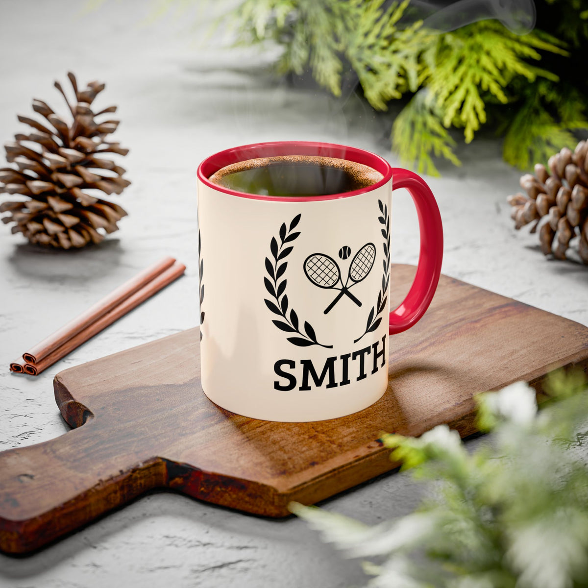 Tennis Coffee Mug A personalized cream-colored ceramic mug with a red handle and rim, featuring a black tennis racket and laurel wreath graphic above the name 'SMITH'. The mug is filled with coffee and sits on a wooden cutting board surrounded by pinecones, cinnamon sticks, and green foliage on a gray stone surface.