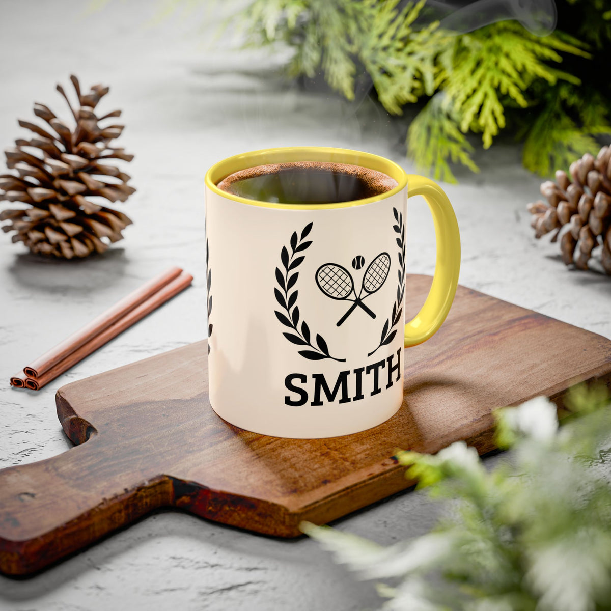 Tennis Coffee Mug A ceramic mug with a yellow handle and rim sits on a wooden cutting board. The mug is cream-colored with a black tennis racket and laurel wreath graphic above the name 'SMITH'.