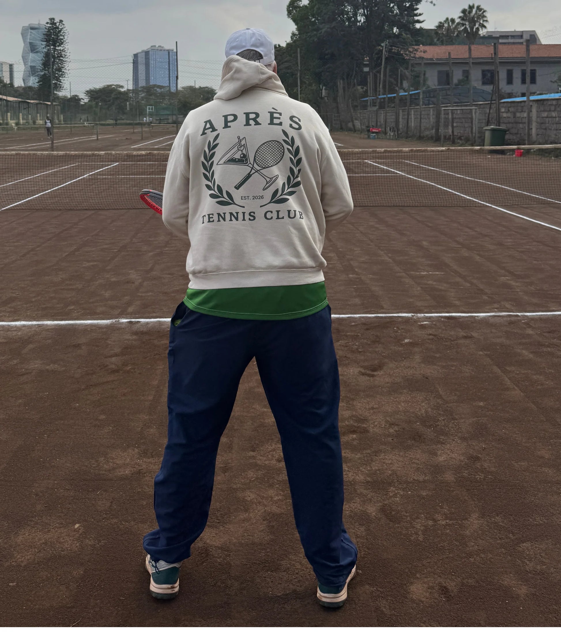 Tennis Hoodie A person standing on a clay tennis court, viewed from behind, wearing an off-white hoodie with a large 'APRÈS TENNIS CLUB' graphic on the back, navy pants, a green undershirt, and a white cap.