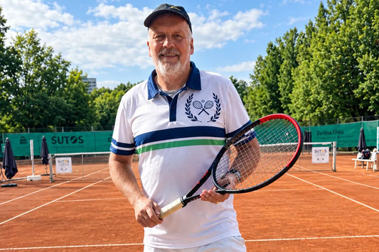 Are_Polos_Appropriate_for_Tennis_Players_ A man wearing a white tennis polo shirt with blue and green stripes, white tennis shorts, and a black cap stands on a clay tennis court holding a racket.