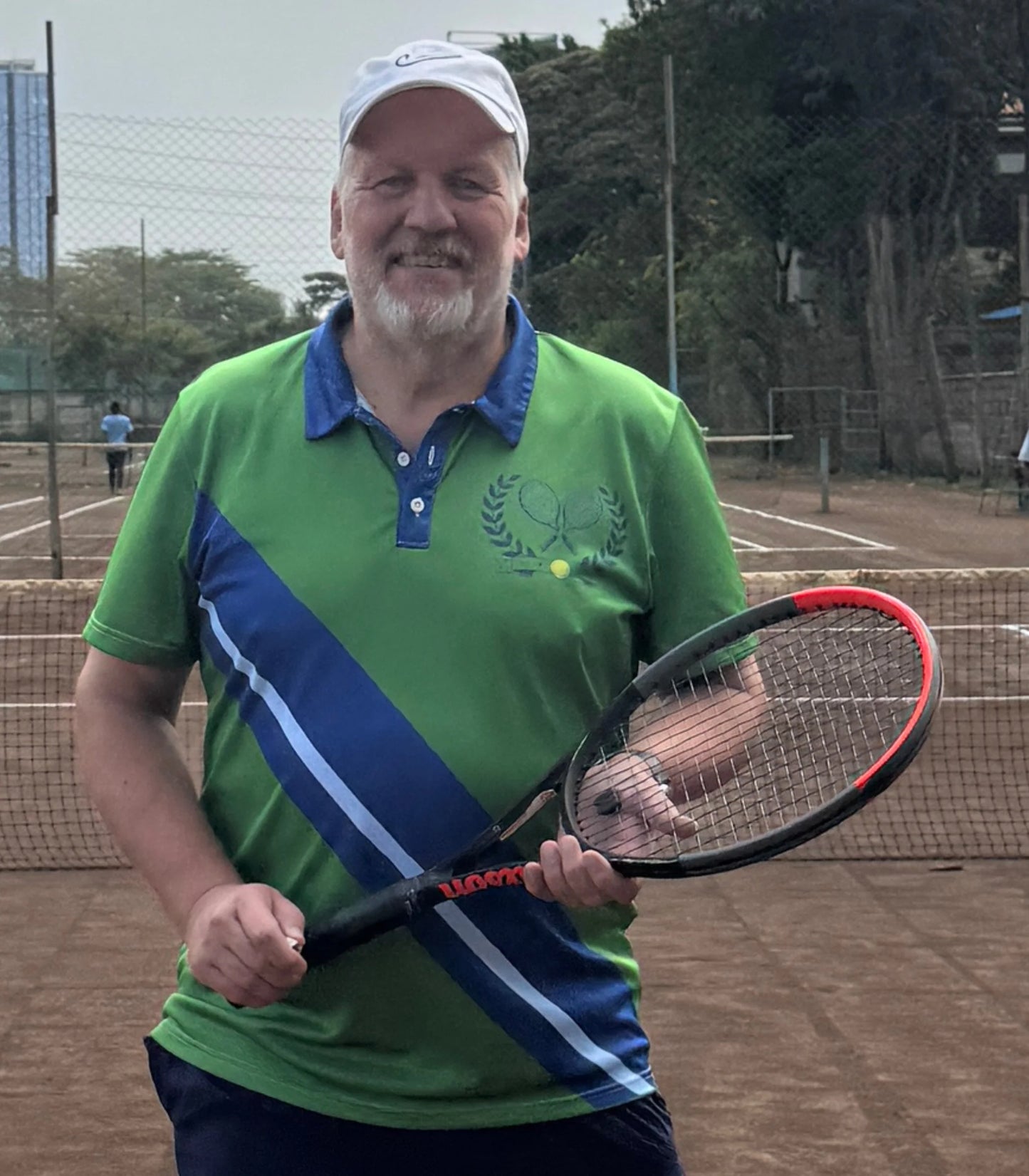 Vintage Tennis Polo A smiling man with a white cap stands on a clay tennis court holding a black and red tennis racket. He is wearing a green and blue polo shirt with a tennis-themed graphic on the chest.