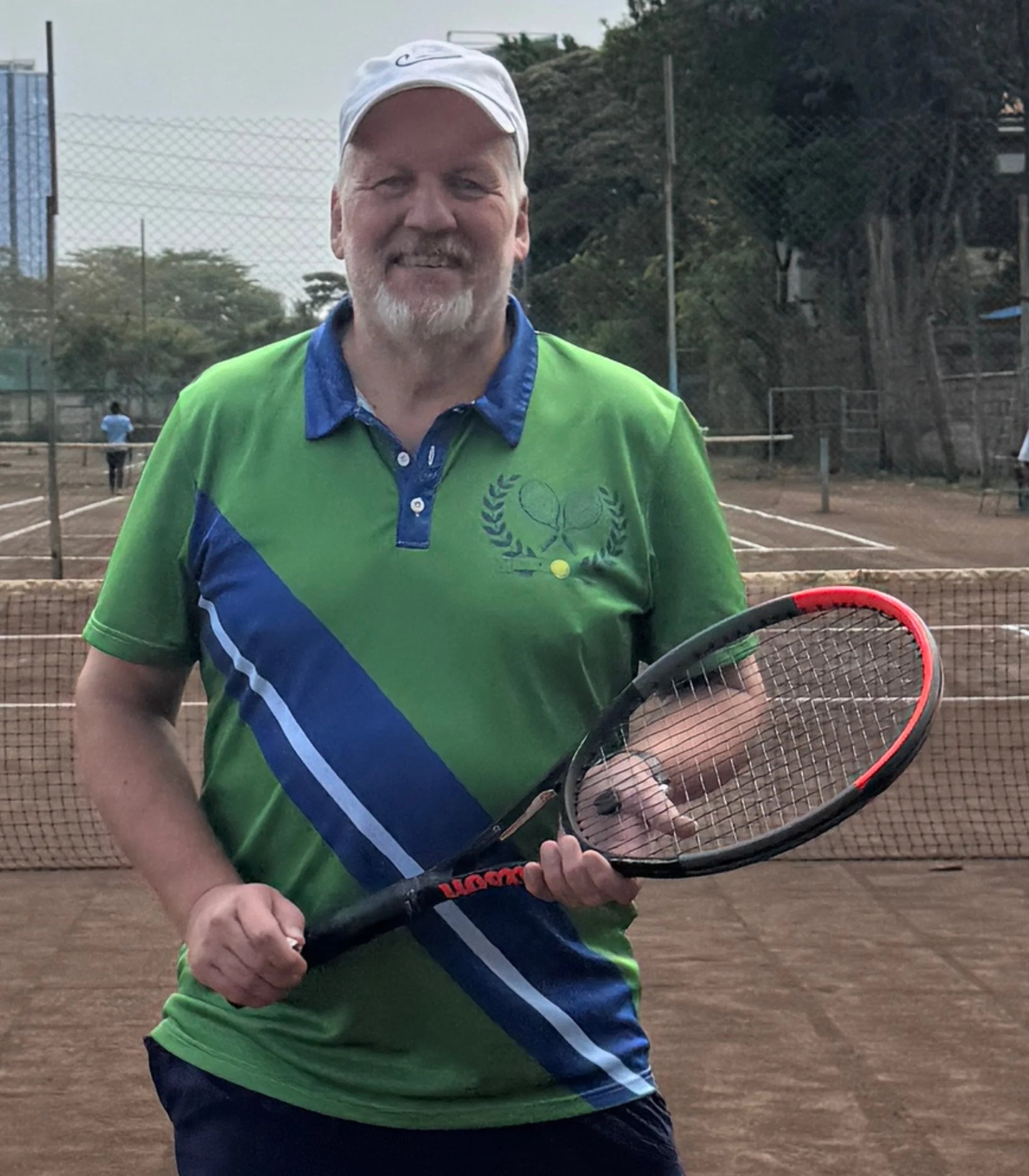 Vintage Tennis Polo A smiling man with a white cap stands on a clay tennis court holding a black and red tennis racket. He is wearing a green and blue polo shirt with a tennis-themed graphic on the chest.