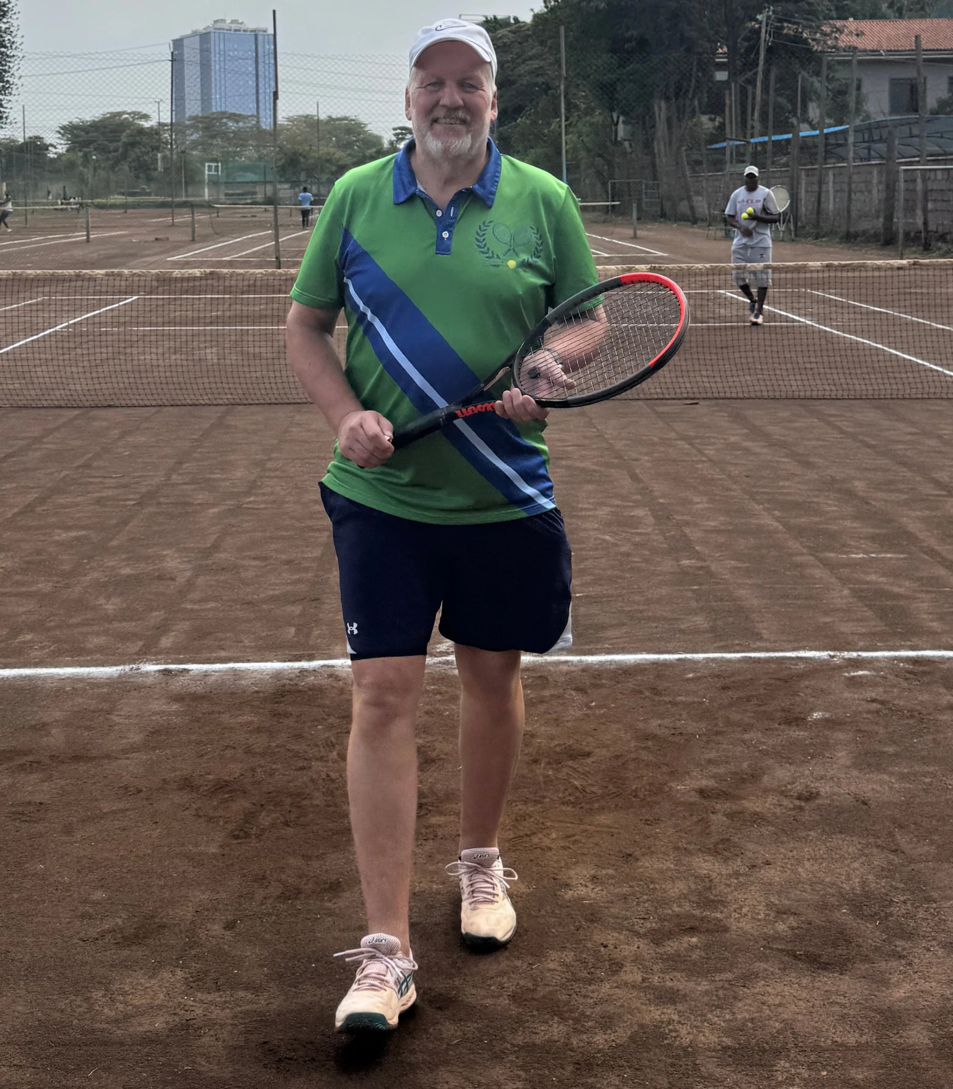 Vintage Tennis Polo A man standing on a clay tennis court holding a tennis racket, wearing a green and blue polo shirt, navy shorts, and white sneakers.