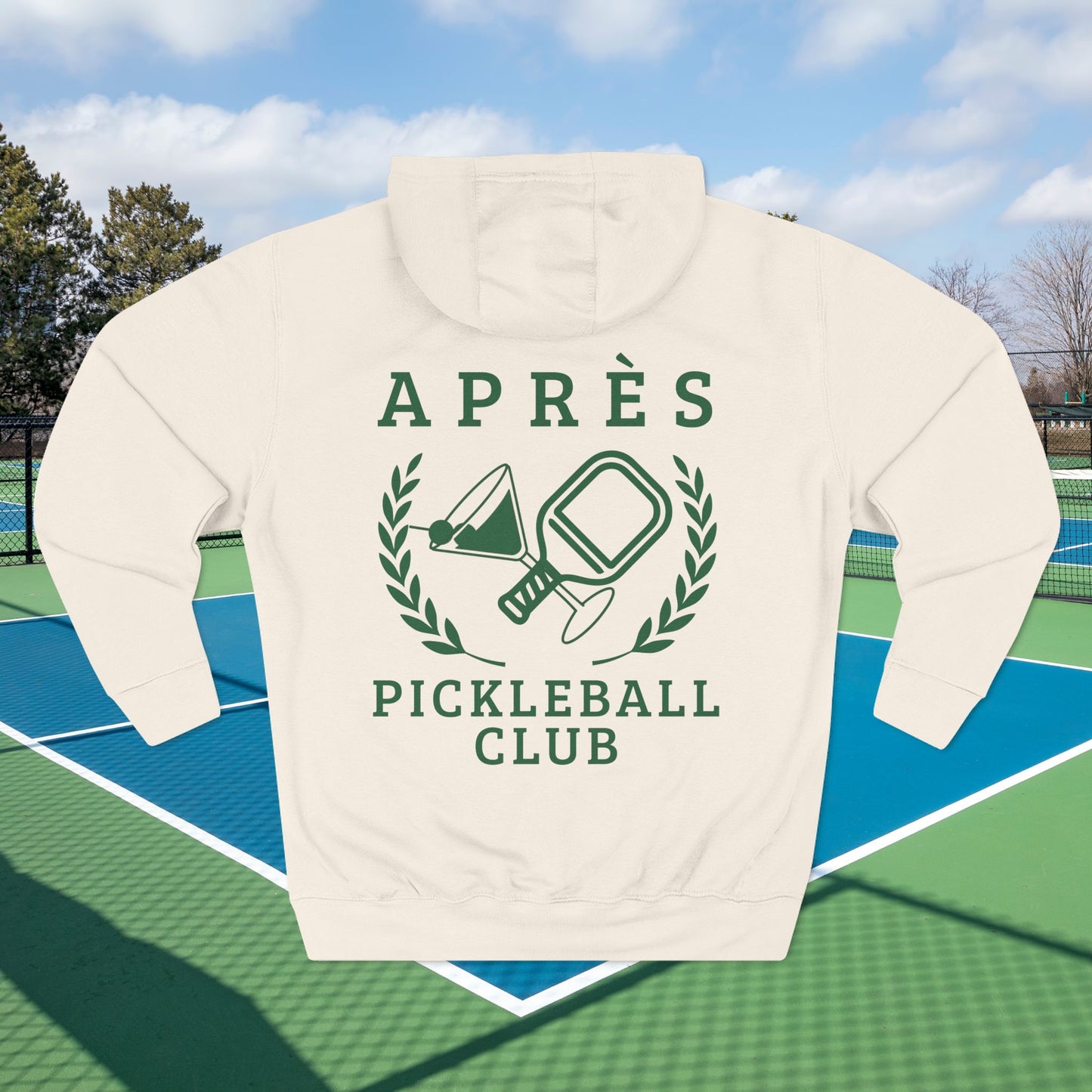 Warm up pickleball hoodie A white pullover hoodie with a green graphic print reading 'APRÈS PICKLEBALL CLUB' and showing a pickleball-themed emblem, displayed on a blue and green pickleball court with a blurred outdoor background.