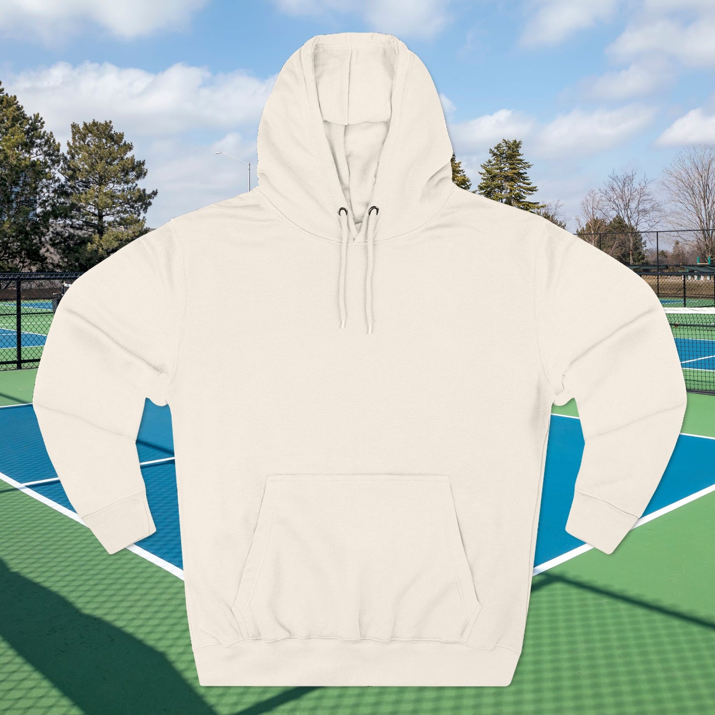 Warm up pickleball hoodie An off-white hooded sweatshirt with a kangaroo pocket and drawstrings, shown against a tennis court background with trees and a blue sky.