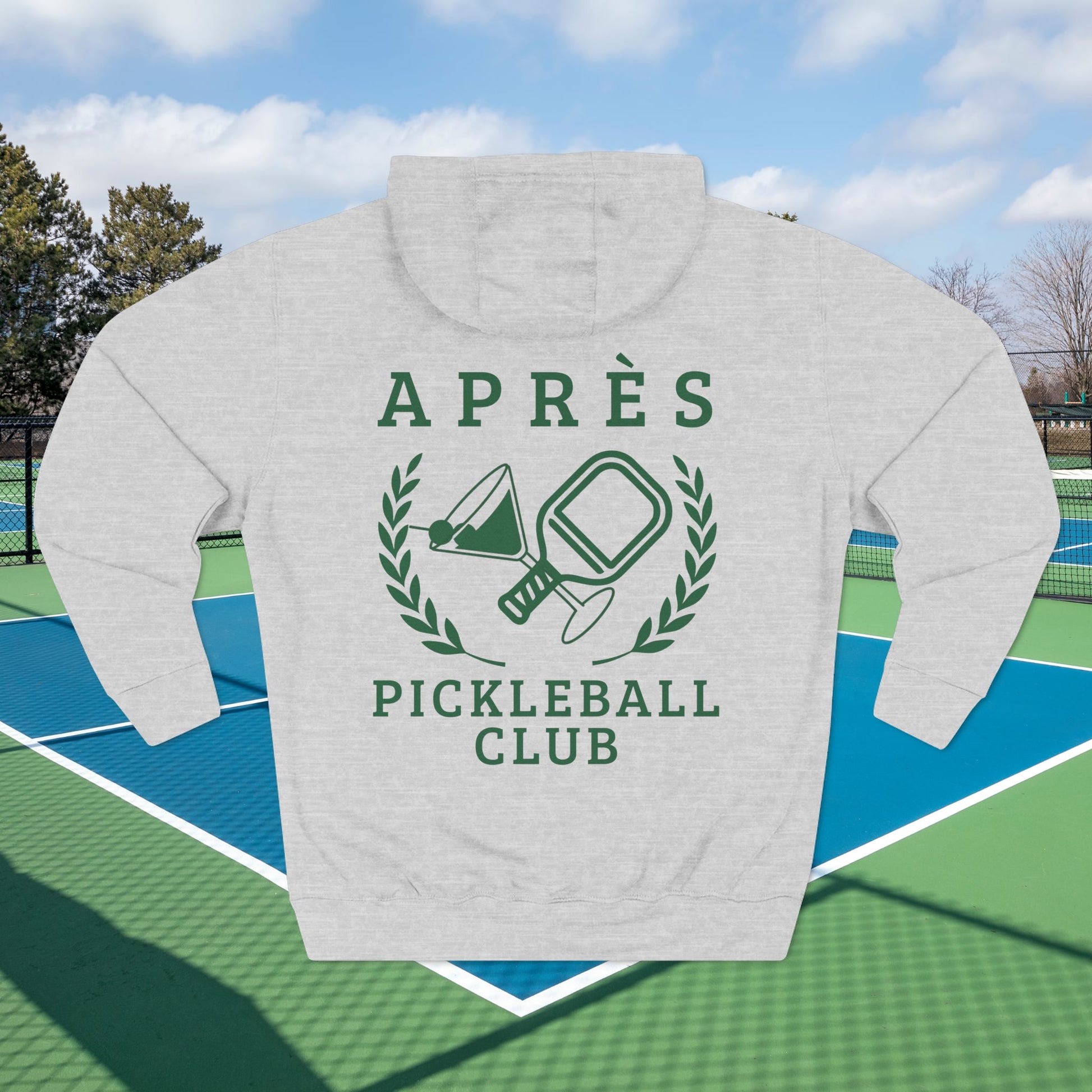 Warm up pickleball hoodie A light heather gray hoodie with a dark green graphic print reading 'APRÈS PICKLEBALL CLUB' and featuring a martini glass and pickleball paddle design, shown as a flat lay on a tennis court with trees and sky in the background.