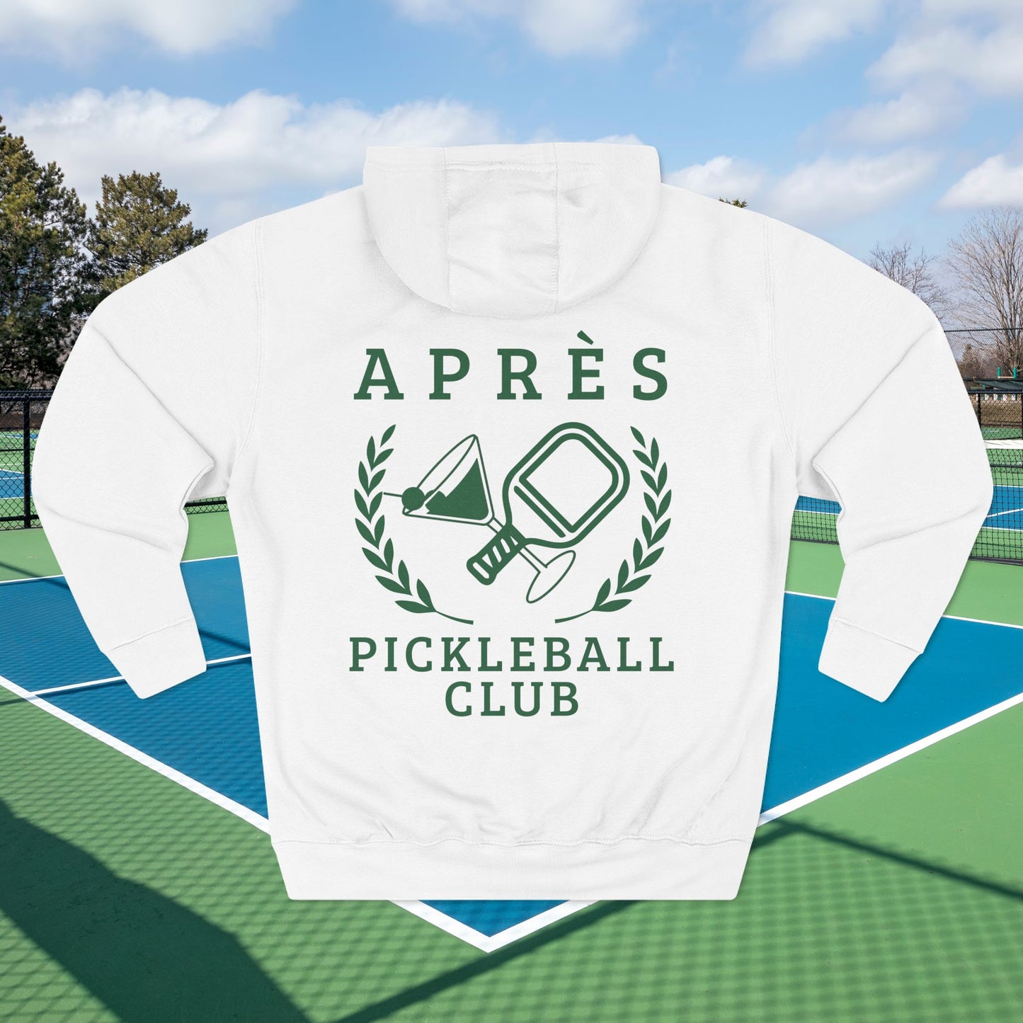 Warm up pickleball hoodie A white pullover hoodie with a green graphic print reading 'APRÈS PICKLEBALL CLUB' and showing a pickleball-themed emblem, displayed on a blue and green pickleball court with a blurred outdoor background.
