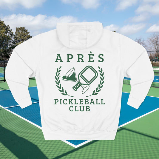 Warm up pickleball hoodie A white pullover hoodie with a green graphic print reading 'APRÈS PICKLEBALL CLUB' and showing a pickleball-themed emblem, displayed on a blue and green pickleball court with a blurred outdoor background.