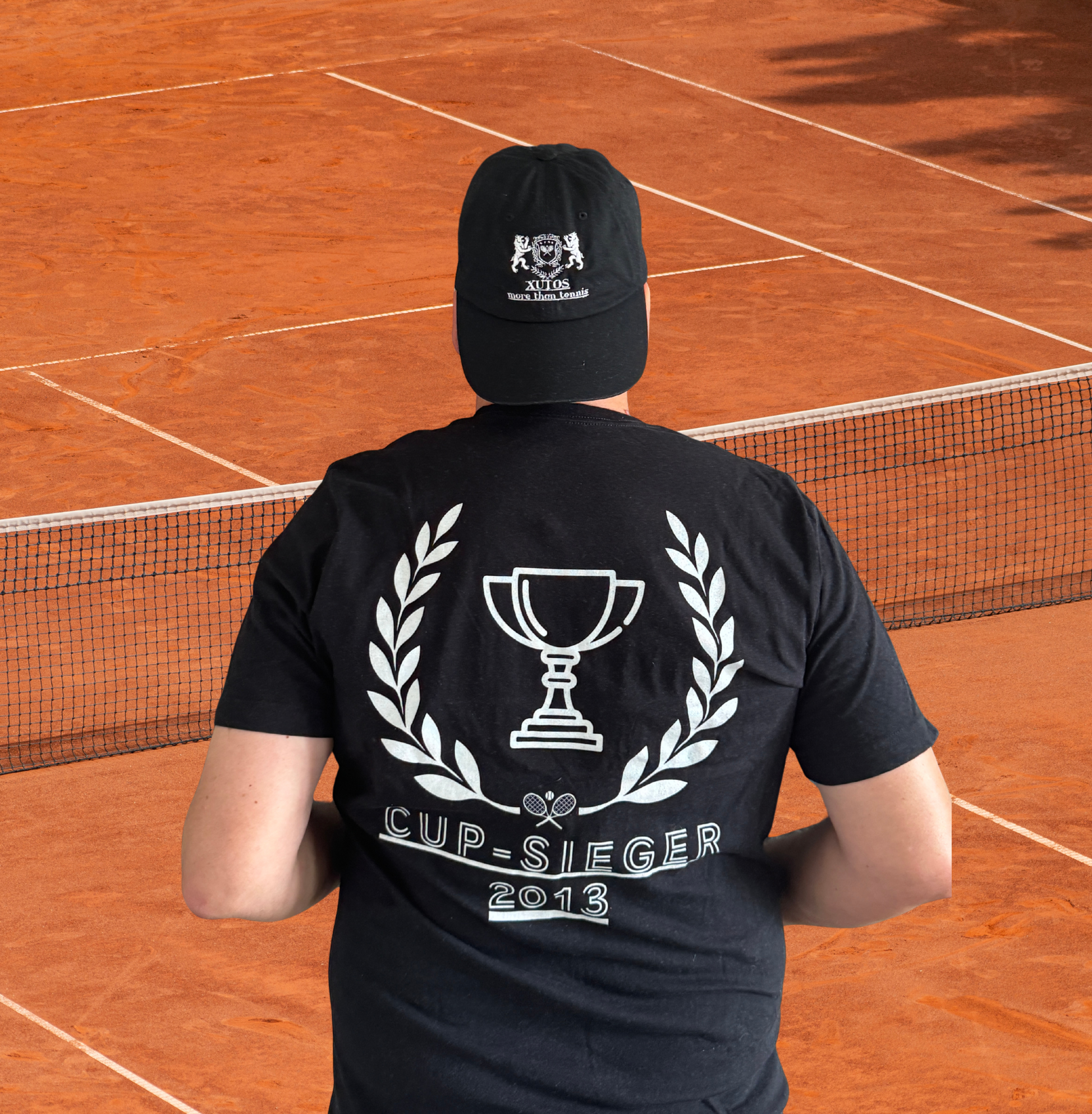A man viewed from behind wearing a black t-shirt and matching black cap on a clay tennis court. The back of the t-shirt features a large white graphic of a trophy framed by laurels with the text 'CUP-SIEGER 2013'.