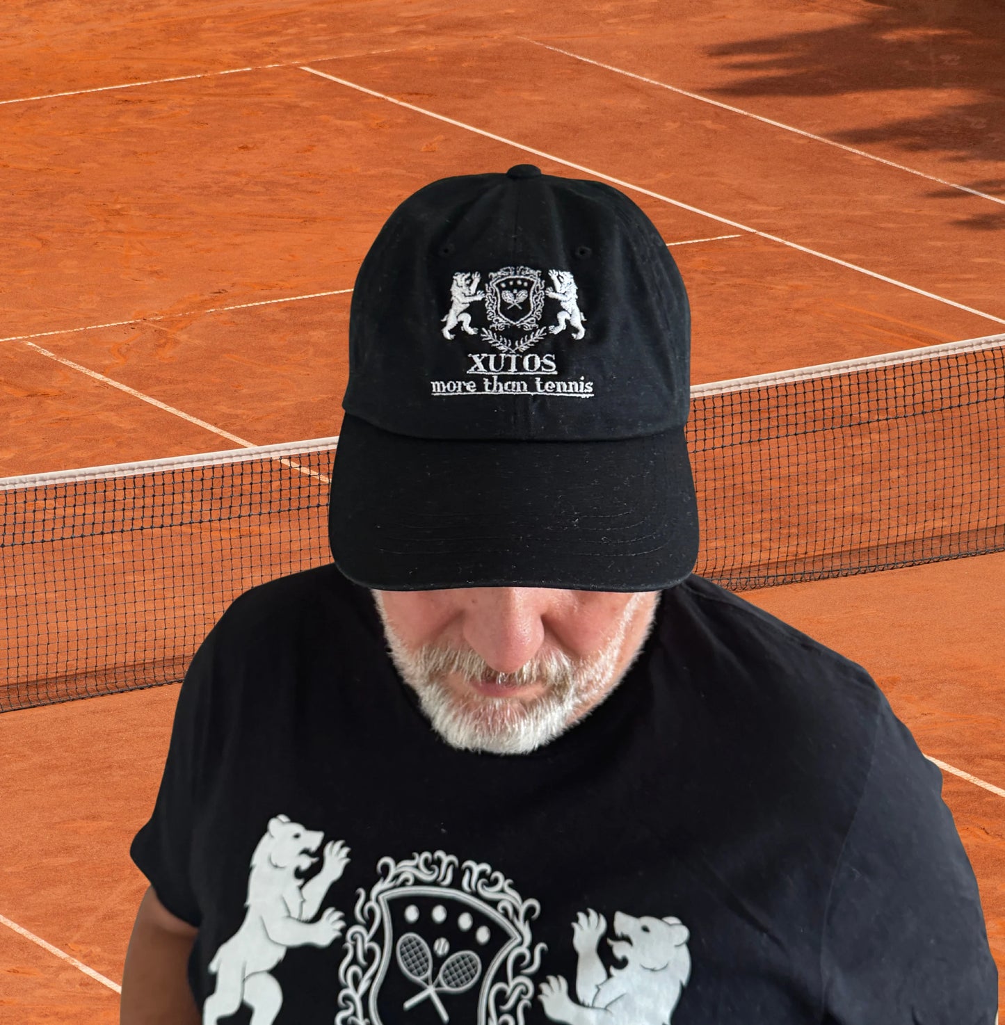 XutosCap A man wearing a black baseball cap with a white embroidered crest and the text 'XULOS' and 'MEXICO CITY' on a clay tennis court.