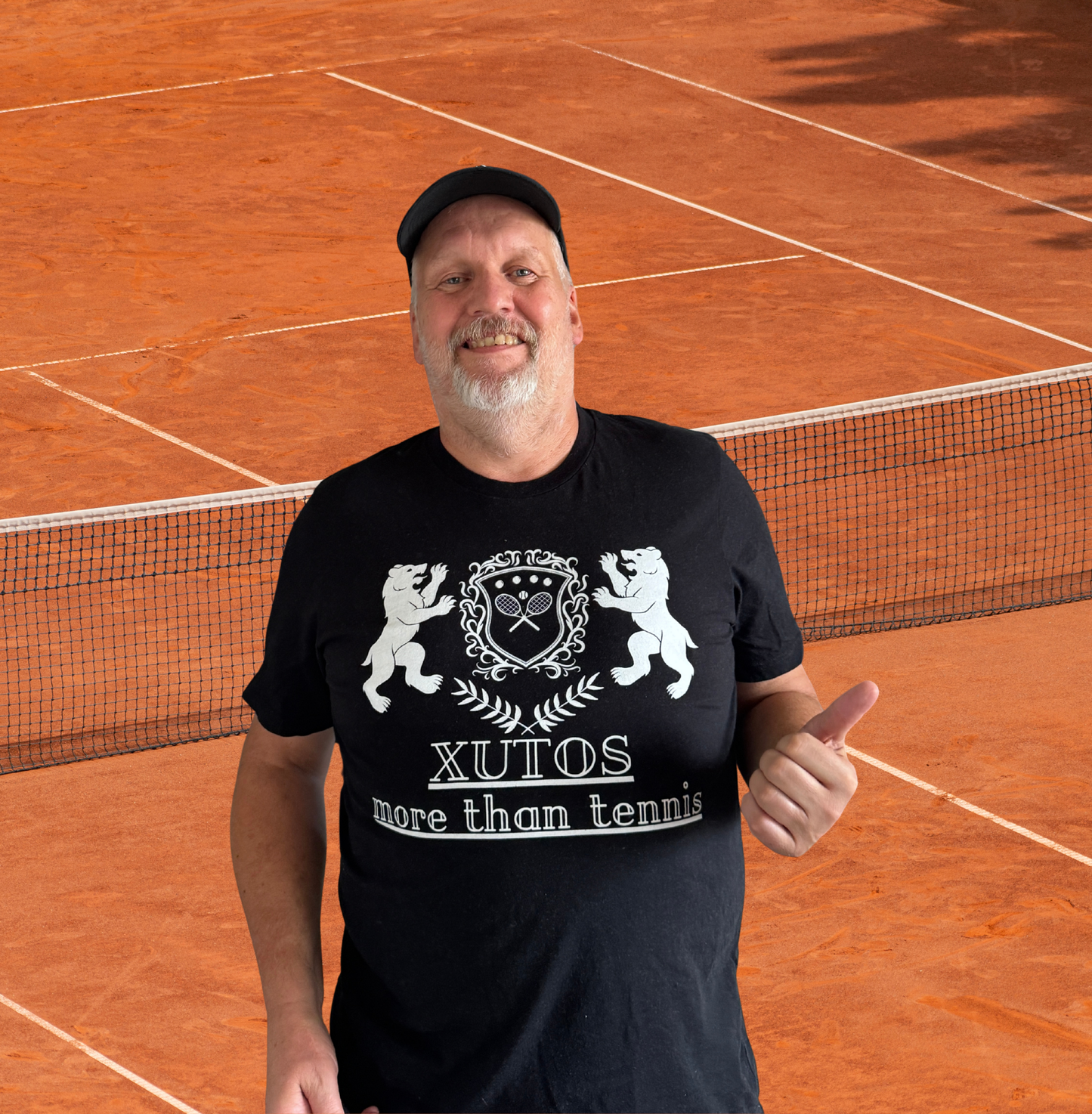 Xutos Polo A man wearing a black t-shirt with a white graphic that reads 'XUTOS more than tennis' stands on a clay tennis court in front of a net, giving a thumbs-up.