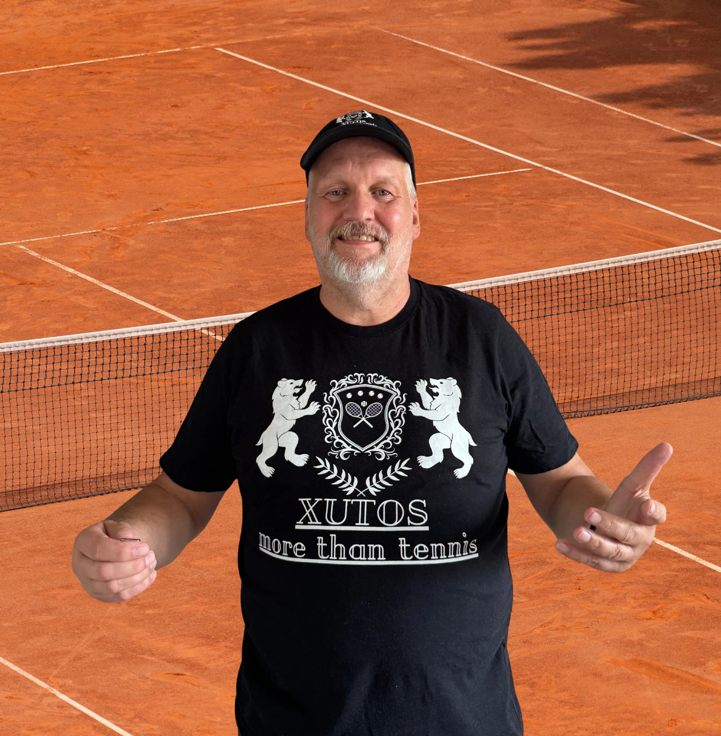 A man wearing a black t-shirt with a white graphic that reads 'XUTOS more than tennis' stands on a clay tennis court. The shirt features a crest with two lions and crossed tennis rackets.