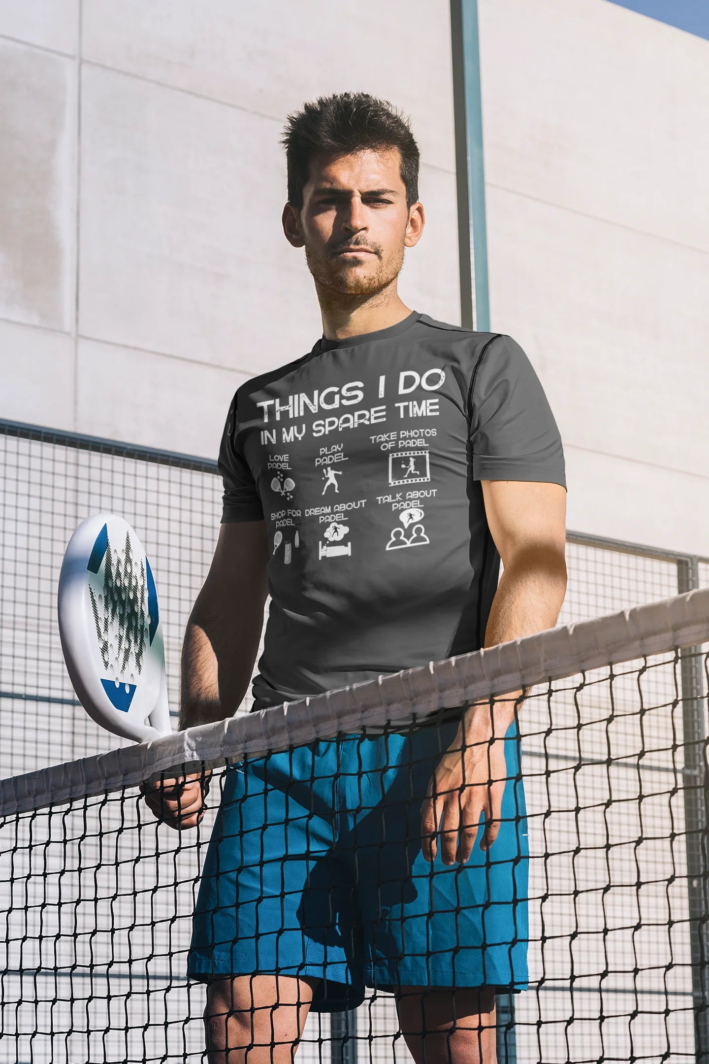 Man holding a paddle on a tennis court wearing a black t-shirt with text and blue shorts.