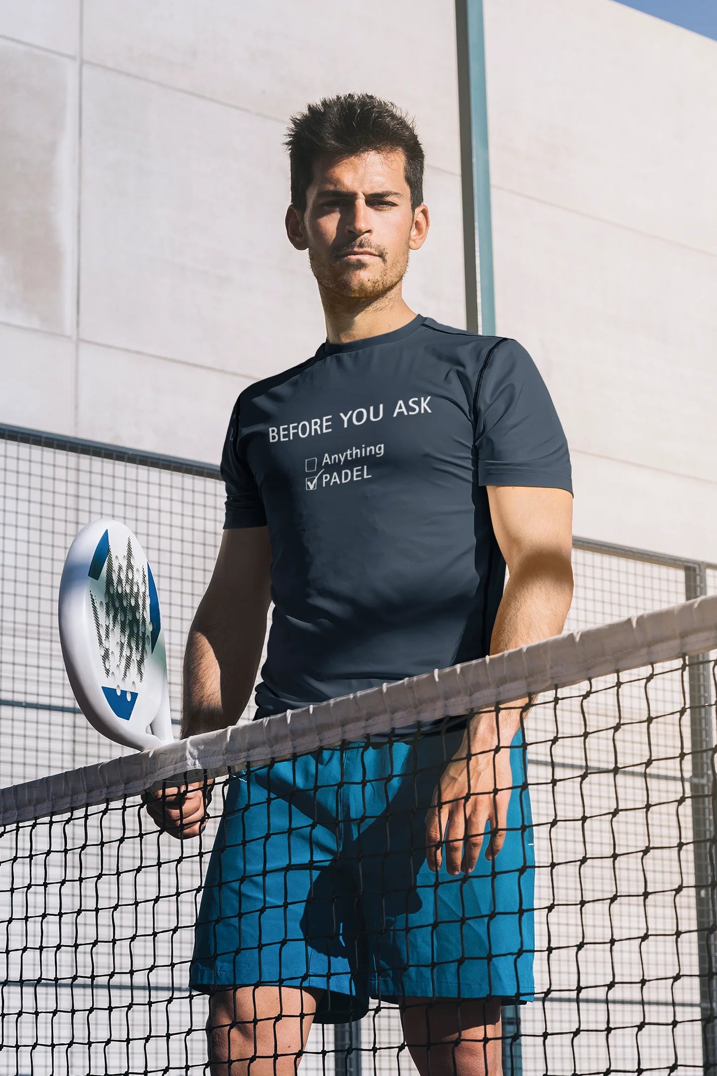 Padel T-Shirt Before you ask Man holding a paddle on a tennis court wearing a branded t-shirt.