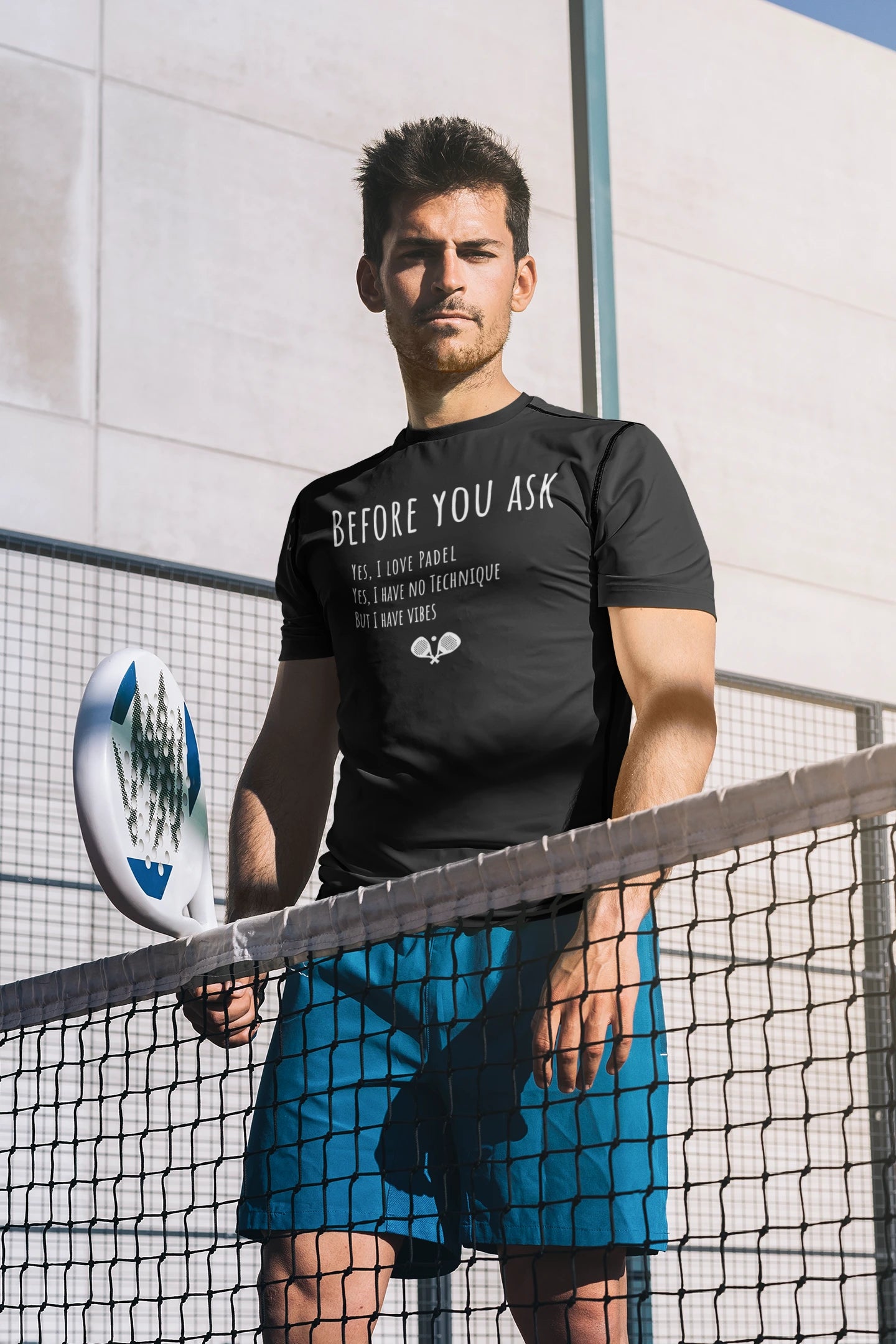Man holding a paddle on a tennis court wearing a black t-shirt with text.