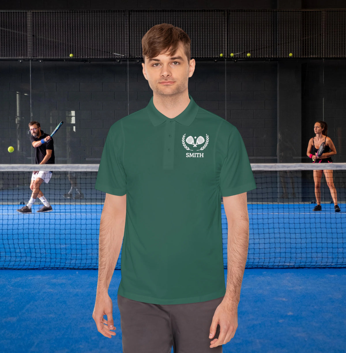 personalized padel gift A man wearing a dark green polo shirt with a white 'SMITH' tennis logo on the chest, standing in front of a tennis court with players in the background.