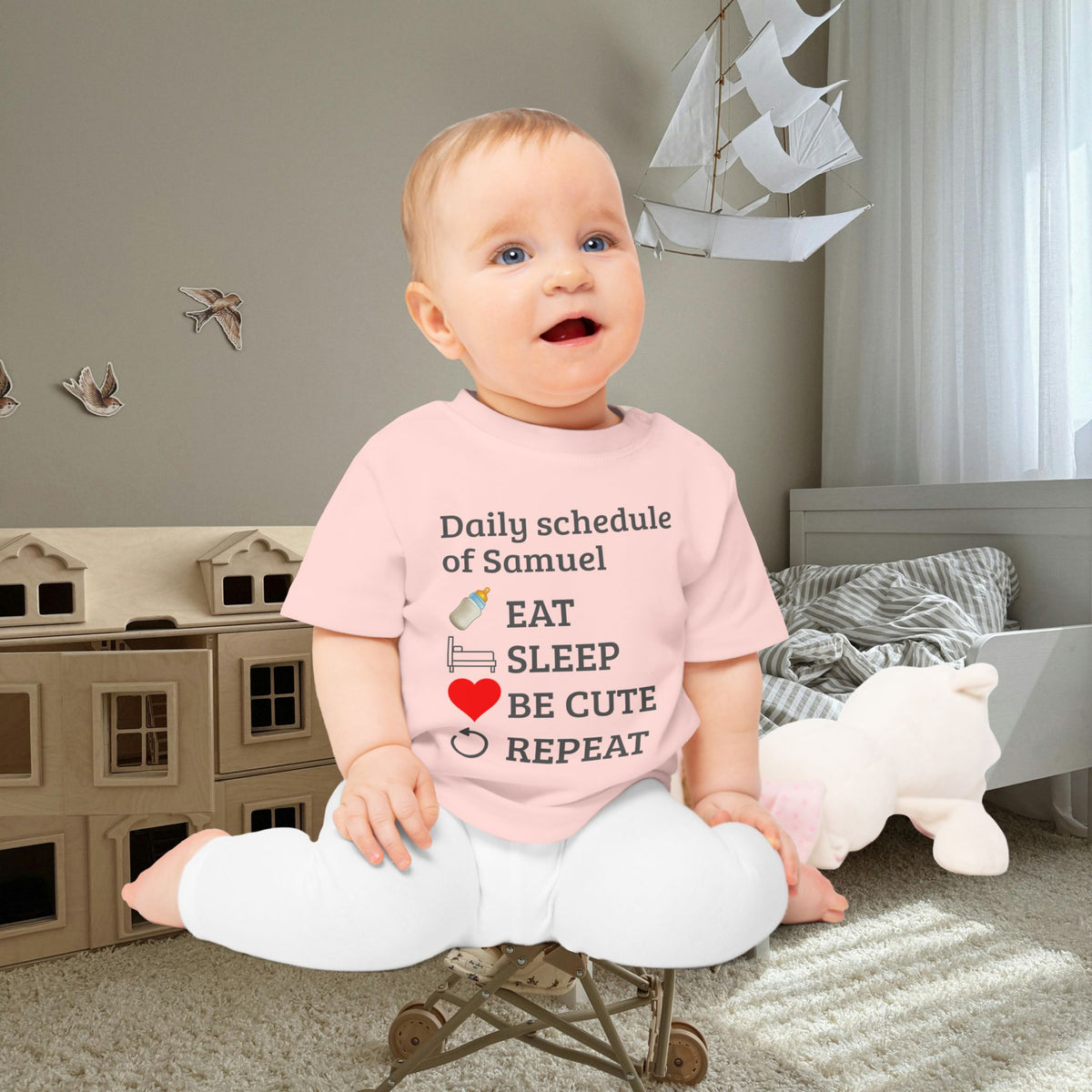 baby gift idea A baby sitting on a small toy cart in a nursery, wearing a light pink graphic t-shirt with the text 'Daily schedule of Samuel EAT SLEEP BE CUTE REPEAT' and white pants.