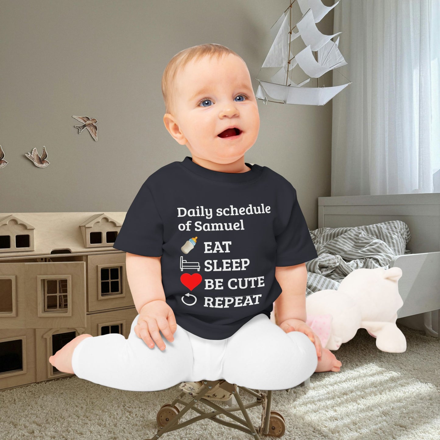 baby gift idea A baby sitting cross-legged in a child's bedroom, wearing a navy blue graphic t-shirt with white text that reads 'Daily schedule of Samuel' followed by an illustration of a bottle, bed, sunglasses, a red heart, and the words 'EAT SLEEP BE CUTE REPEAT'.