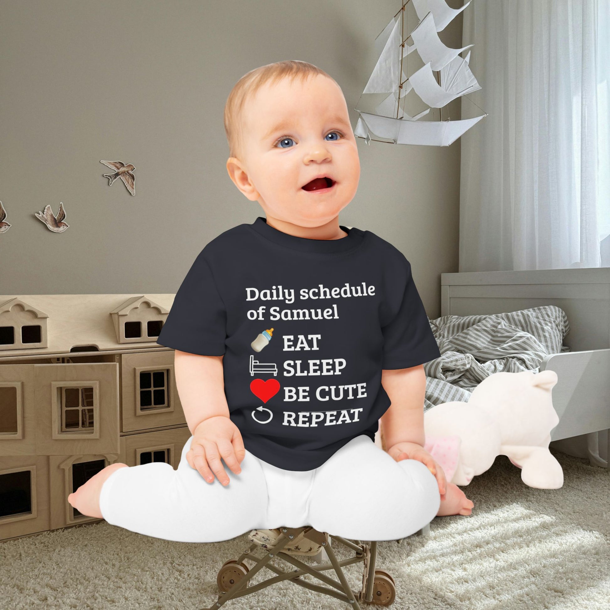baby gift idea A baby sitting cross-legged in a child's bedroom, wearing a navy blue graphic t-shirt with white text that reads 'Daily schedule of Samuel' followed by an illustration of a bottle, bed, sunglasses, a red heart, and the words 'EAT SLEEP BE CUTE REPEAT'.
