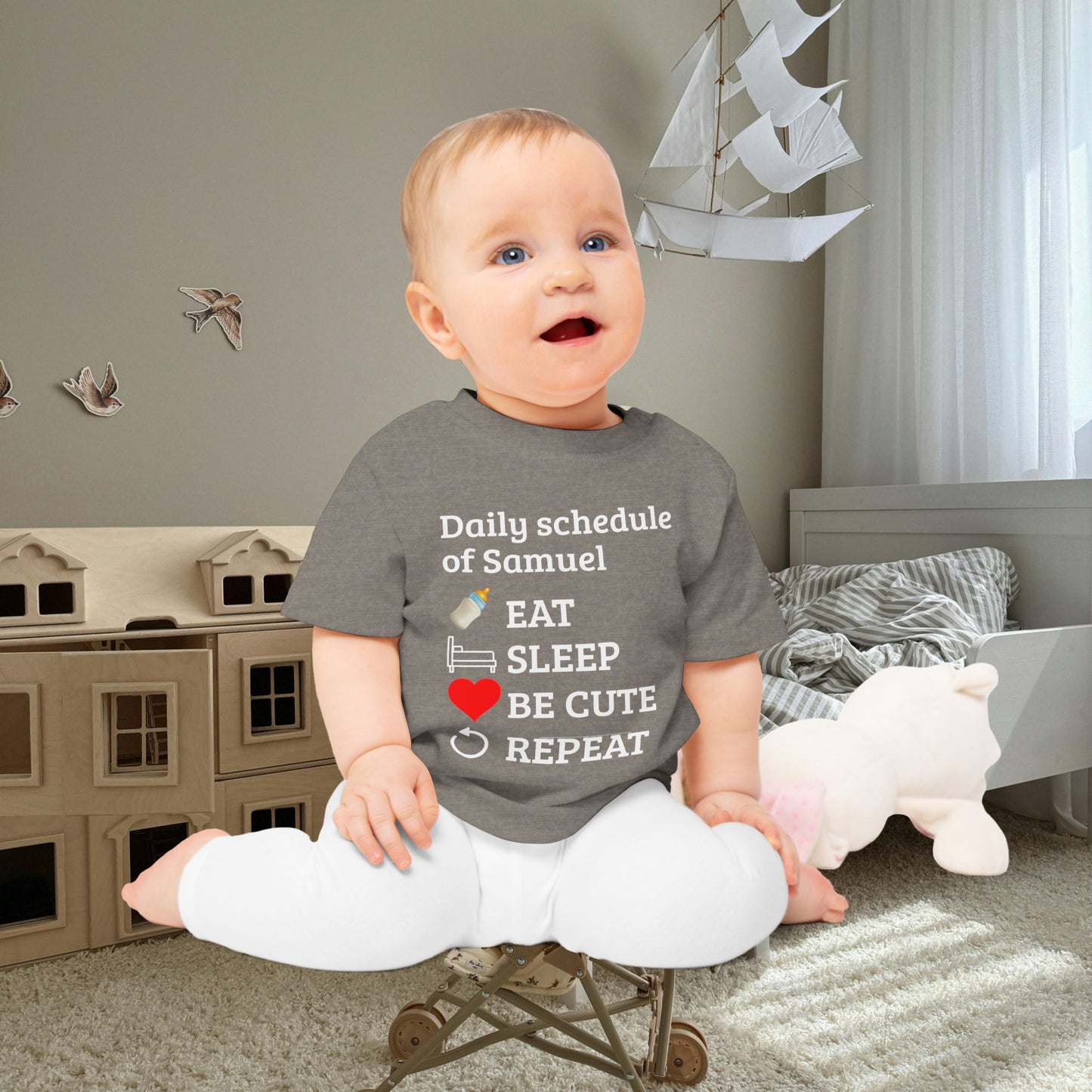 baby gift idea A baby sitting on a small toy chair in a child's bedroom, wearing a dark gray graphic t-shirt with white text and illustrations that reads 'Daily schedule of Samuel EAT SLEEP BE CUTE REPEAT' with small icons for a bottle, pacifier, and heart.