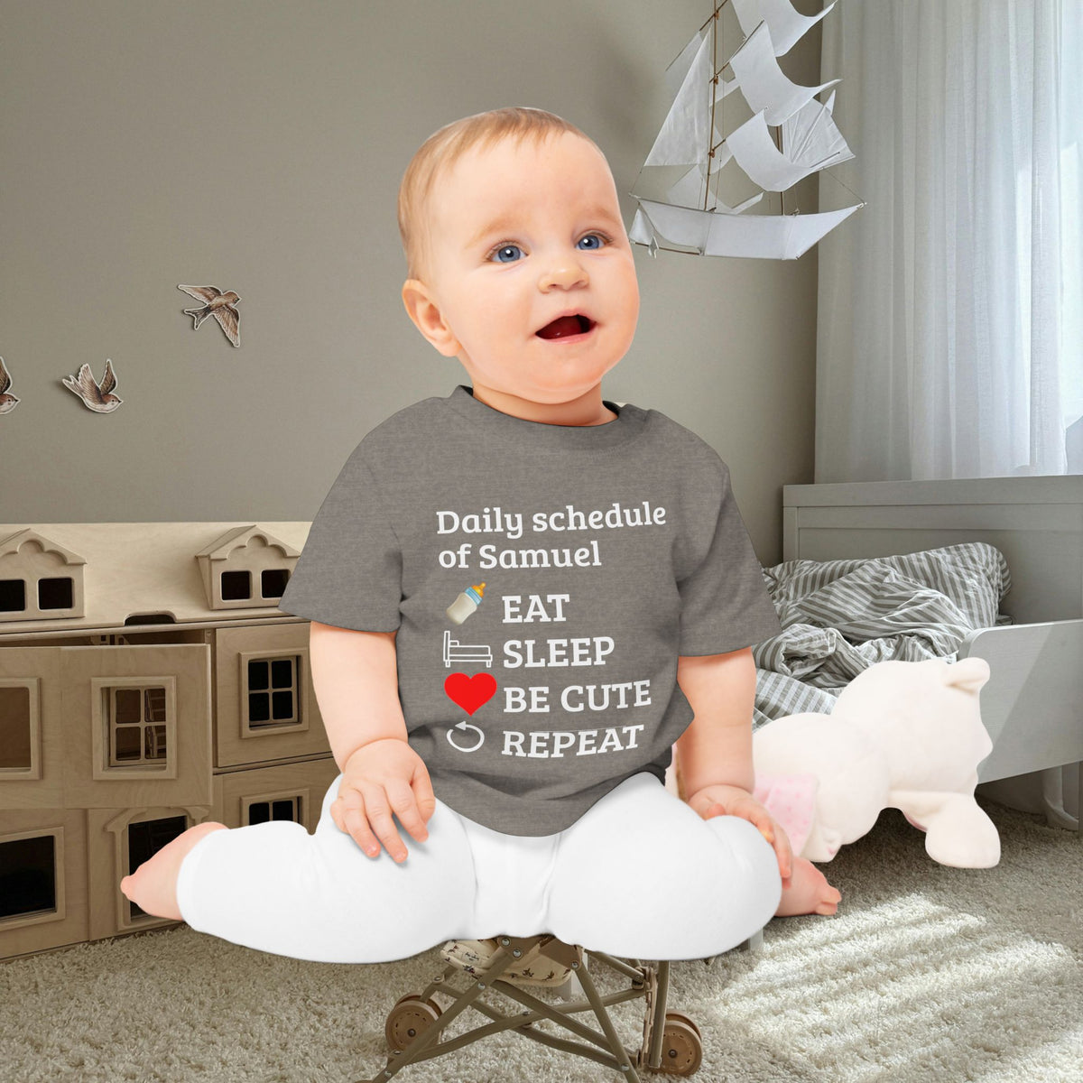 baby gift idea A baby sitting on a small toy chair in a child's bedroom, wearing a dark gray graphic t-shirt with white text and illustrations that reads 'Daily schedule of Samuel EAT SLEEP BE CUTE REPEAT' with small icons for a bottle, pacifier, and heart.