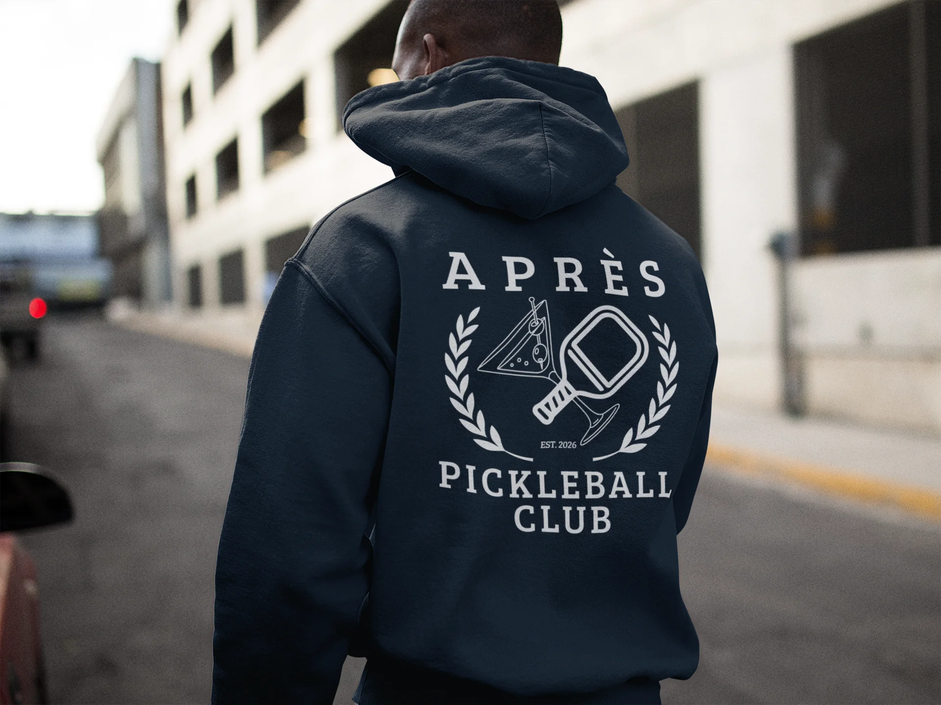 Funny Pickleball Hoodie Person wearing a navy hoodie with 'APRèS Pickleball Club' design on the back, standing on a street.