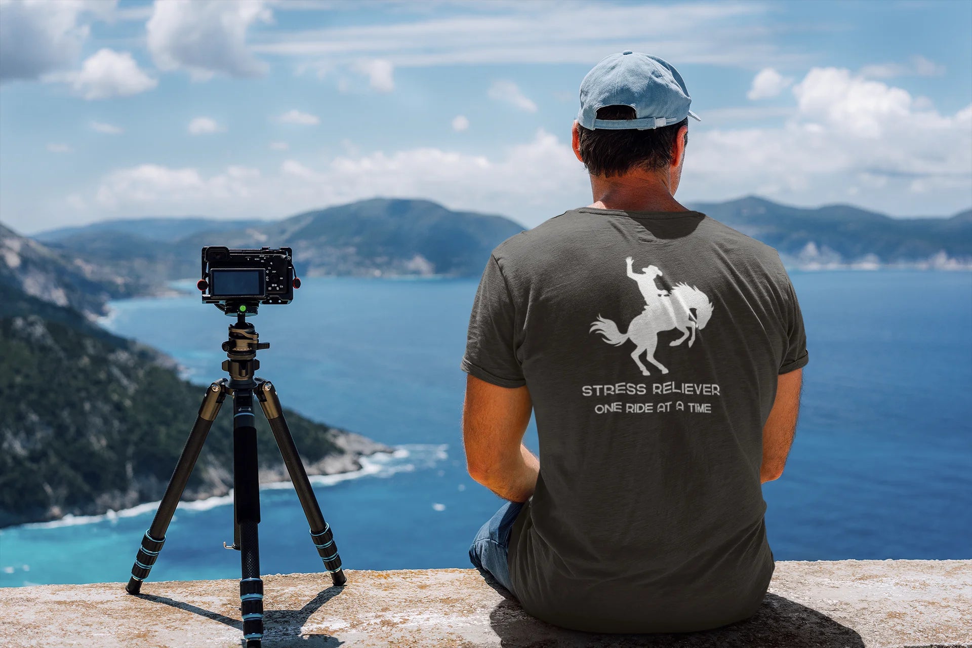 Person sitting on a ledge with a camera on a tripod overlooking a scenic view of mountains and water.