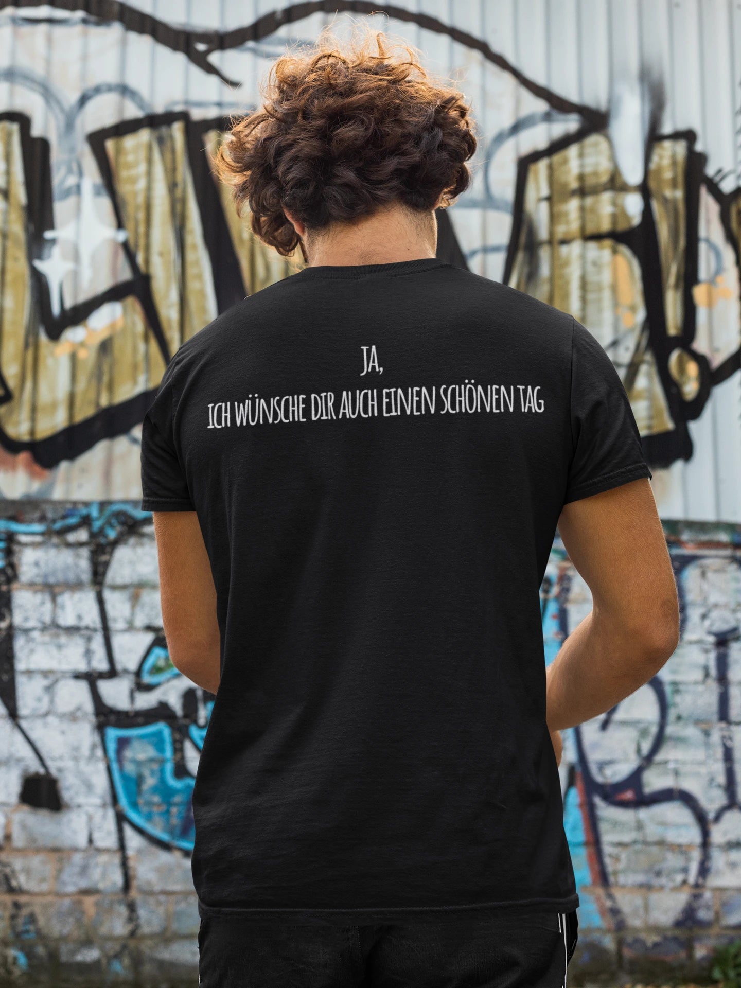Person wearing a black t-shirt with text on the back in front of a graffiti-covered wall.