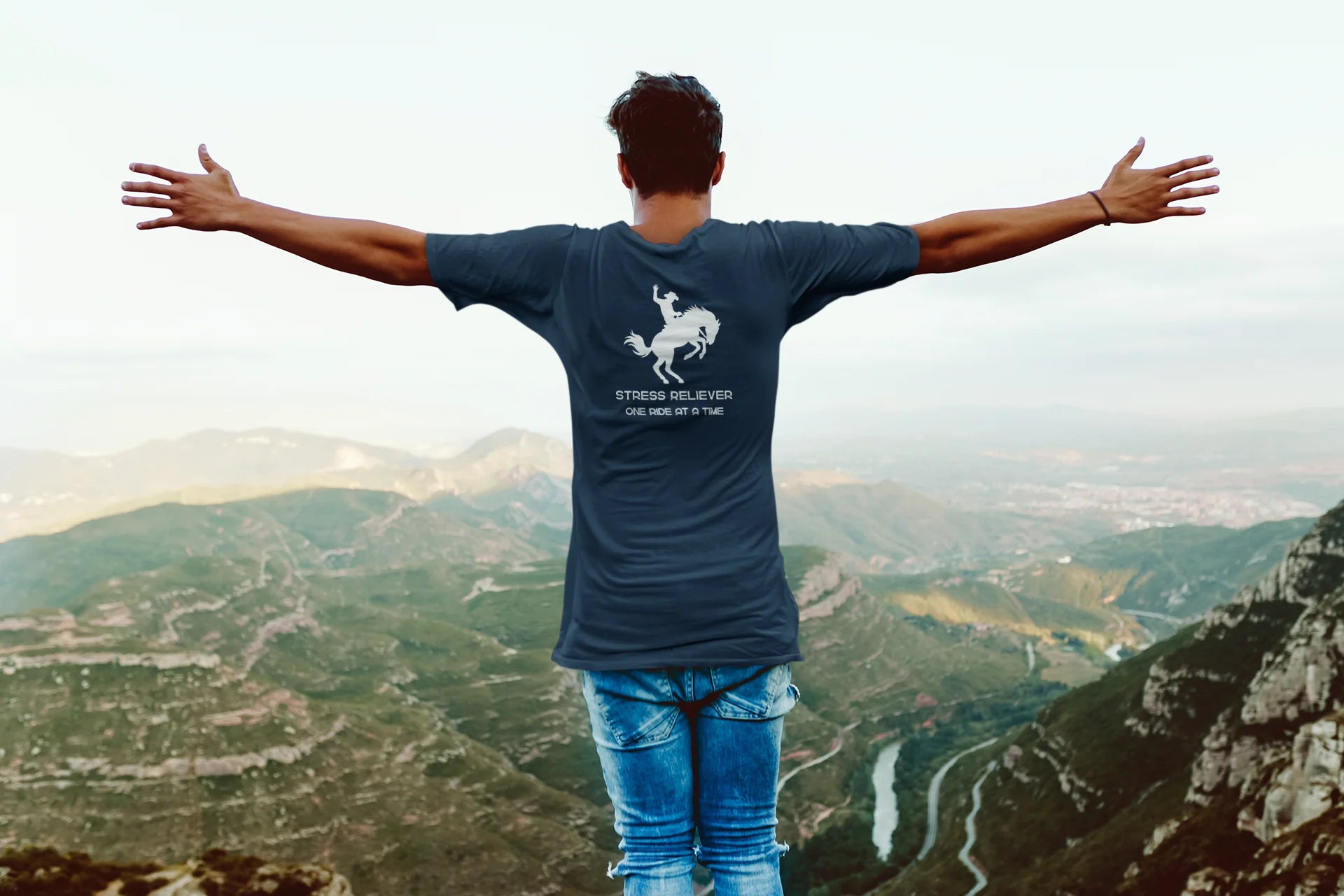 Person wearing a blue t-shirt with a horse graphic and text, standing on a mountain with a scenic view.