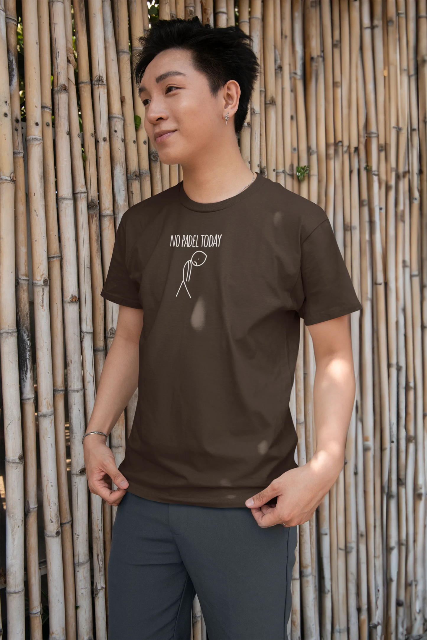 Person wearing a brown t-shirt with a graphic design against a bamboo wall.