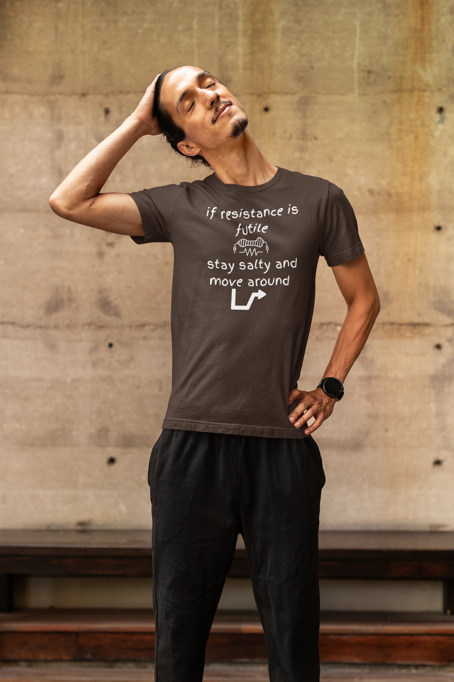 Man wearing a brown t-shirt with text and black pants against a concrete wall.