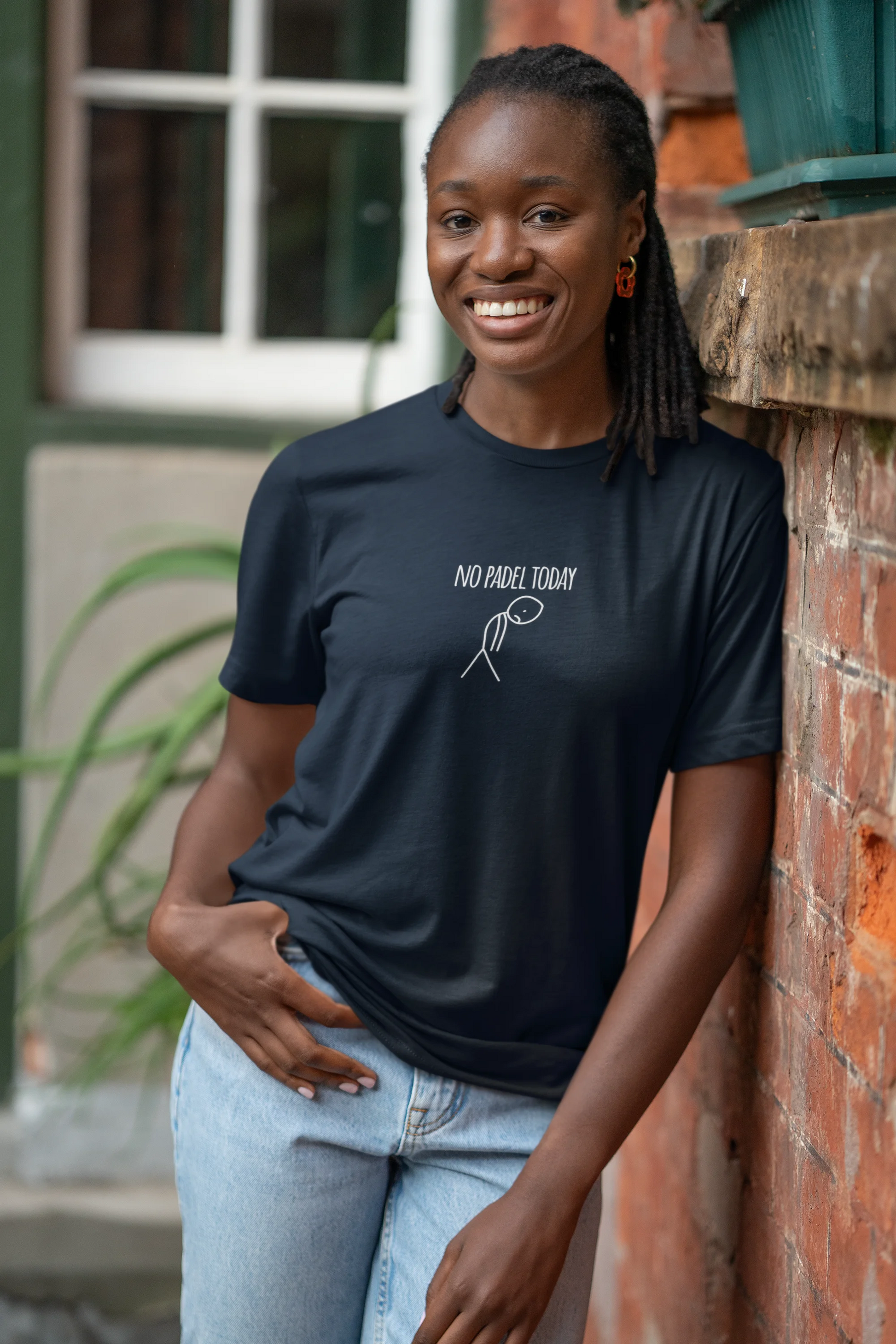 Woman wearing a black t-shirt with text and a logo, standing against a brick wall.