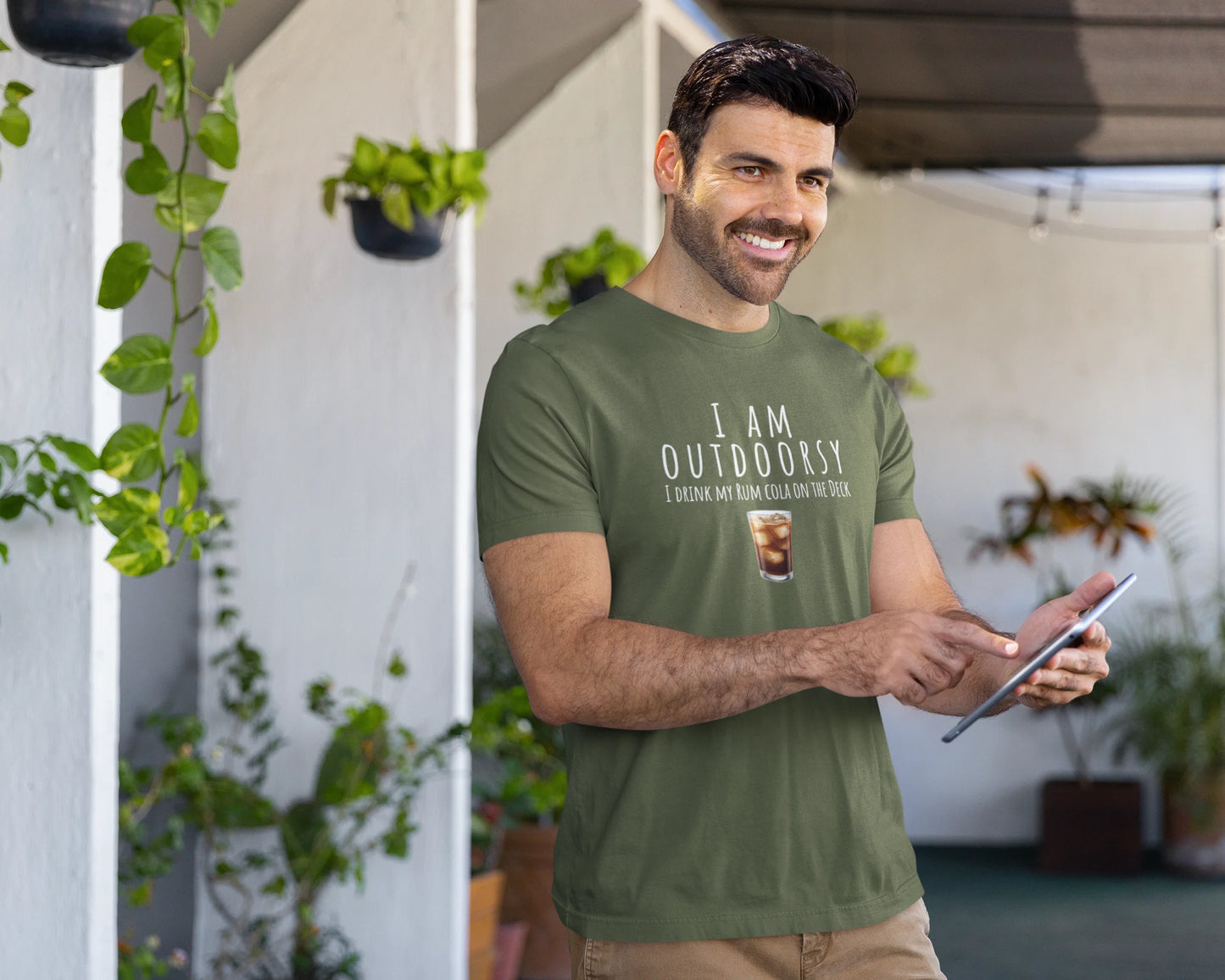 Man wearing a green t-shirt with 'I AM OUTDOORSY' text, holding a tablet, standing in an outdoor setting with plants.