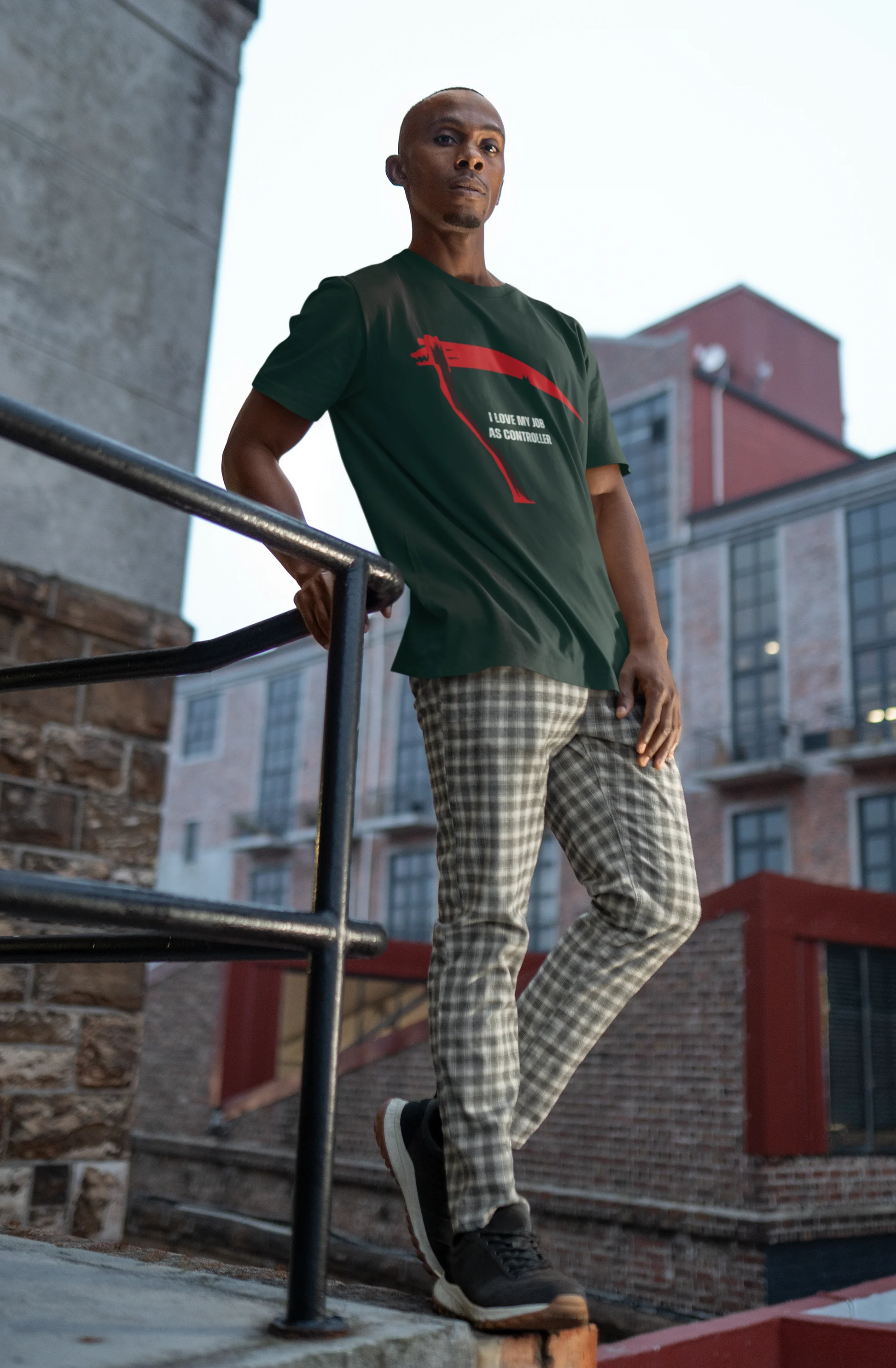 Man wearing a green t-shirt with a red graphic design, standing on a ledge with urban buildings in the background.