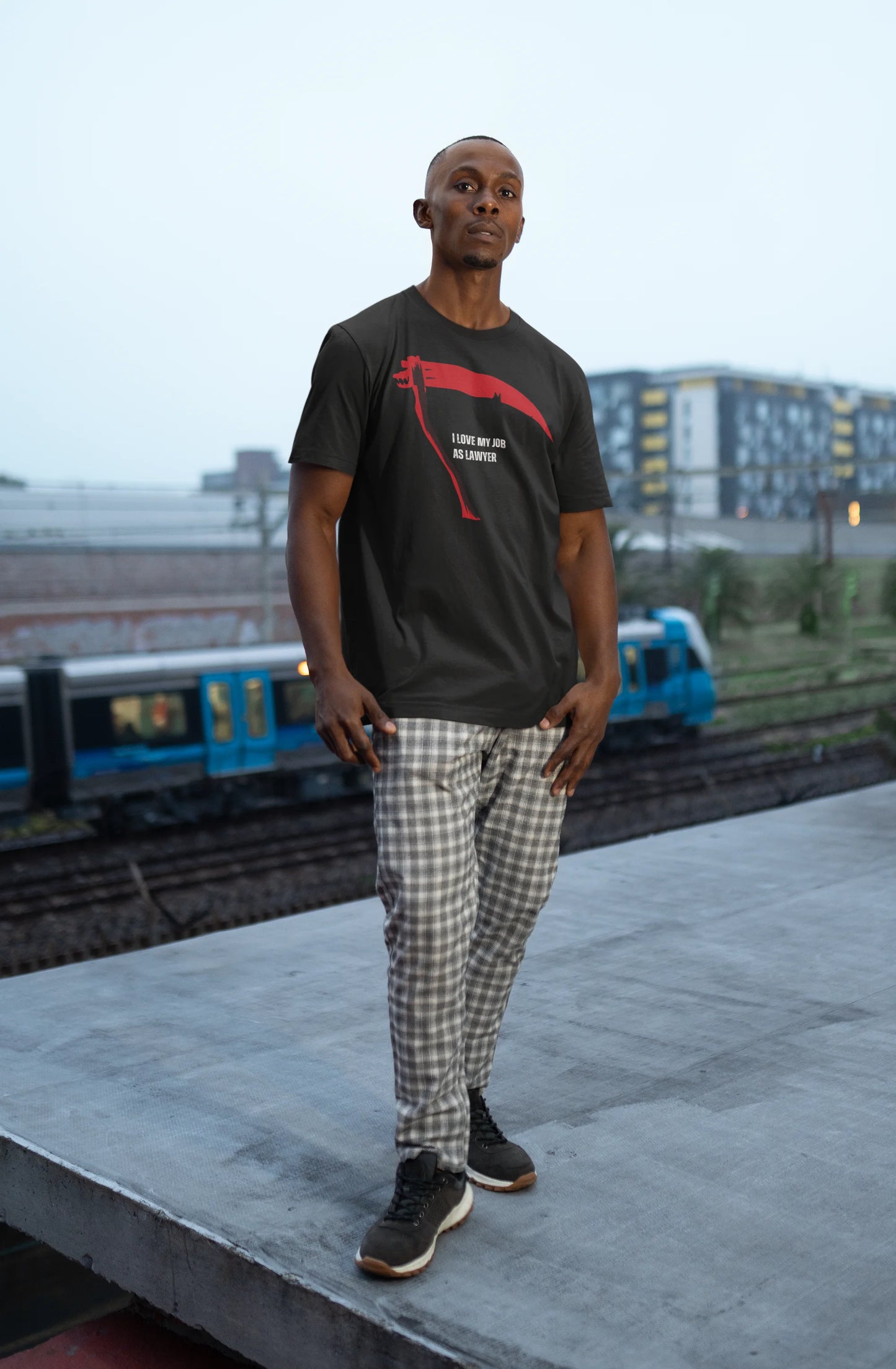 Man wearing a black t-shirt with red design and plaid pants standing on a platform with trains in the background.