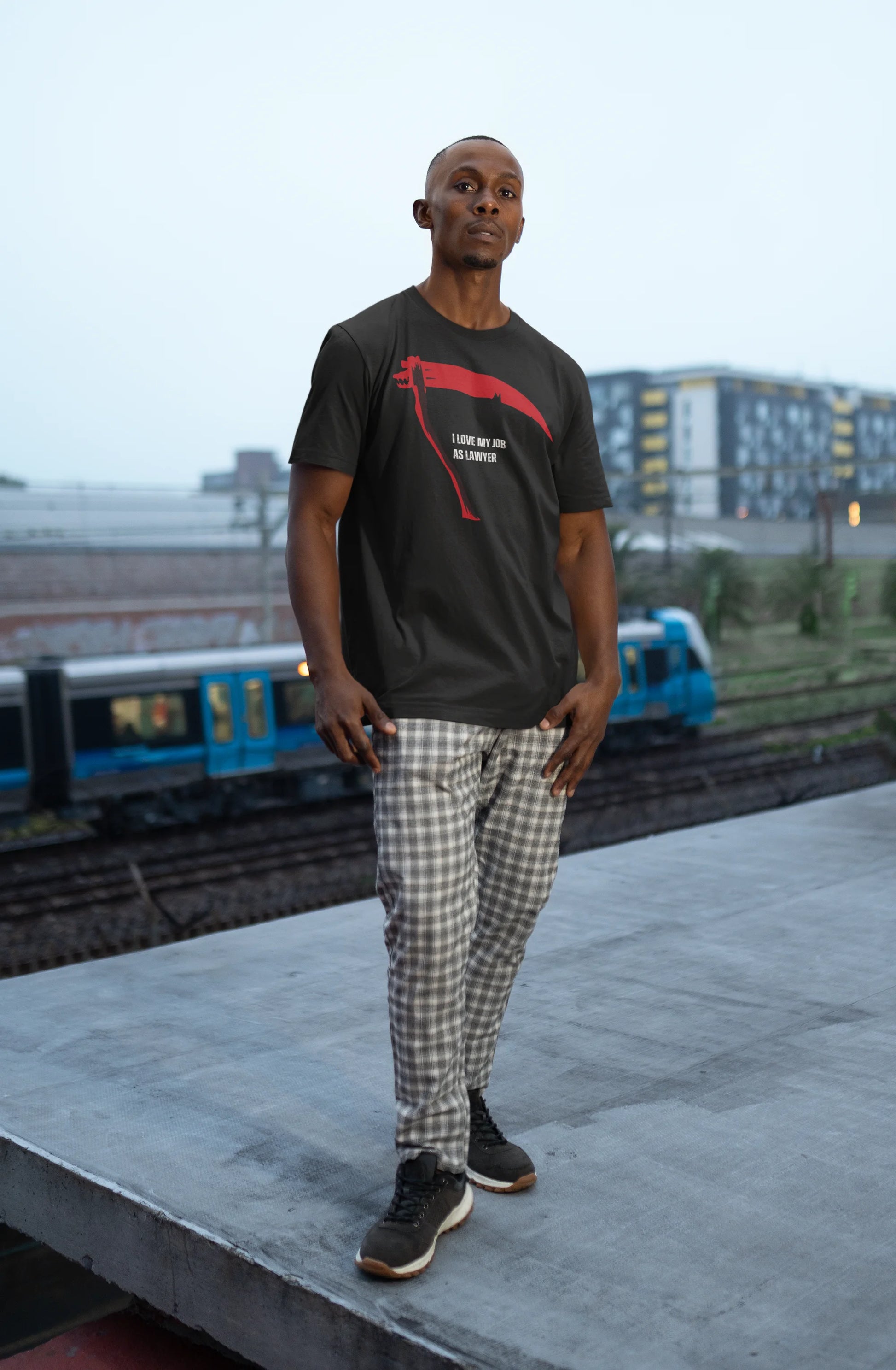 Funny Lawyer Shirt Man wearing a black t-shirt with red design and plaid pants standing on a platform with trains in the background.