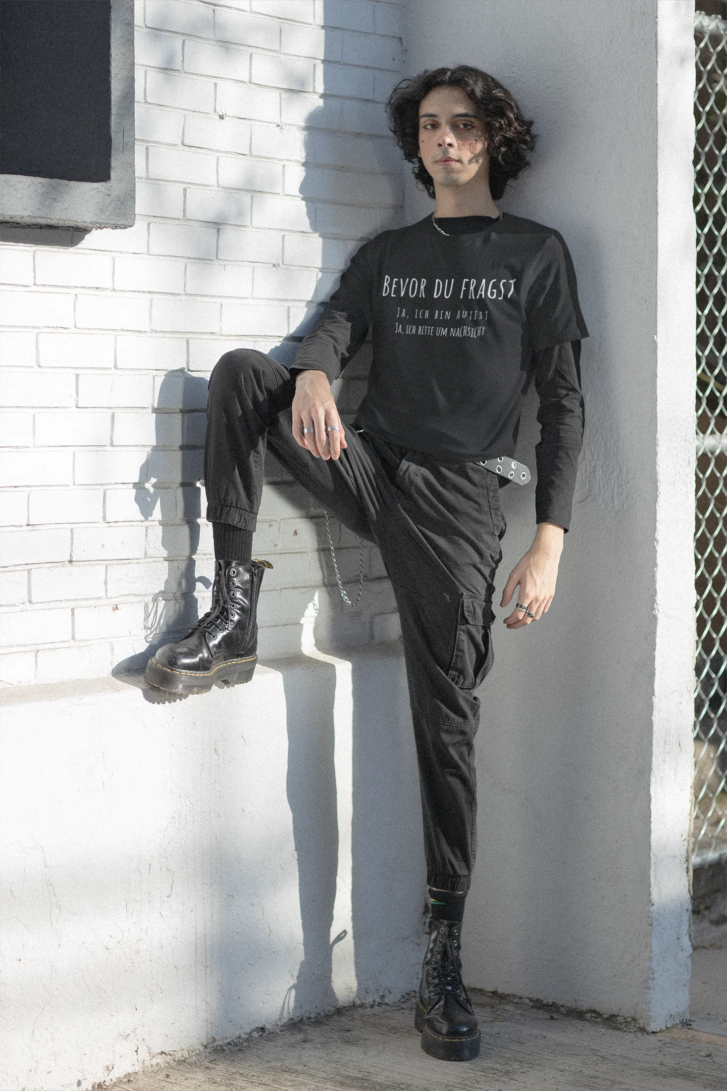 Person wearing a black sweatshirt with text, black pants, and black boots against a white brick wall.
