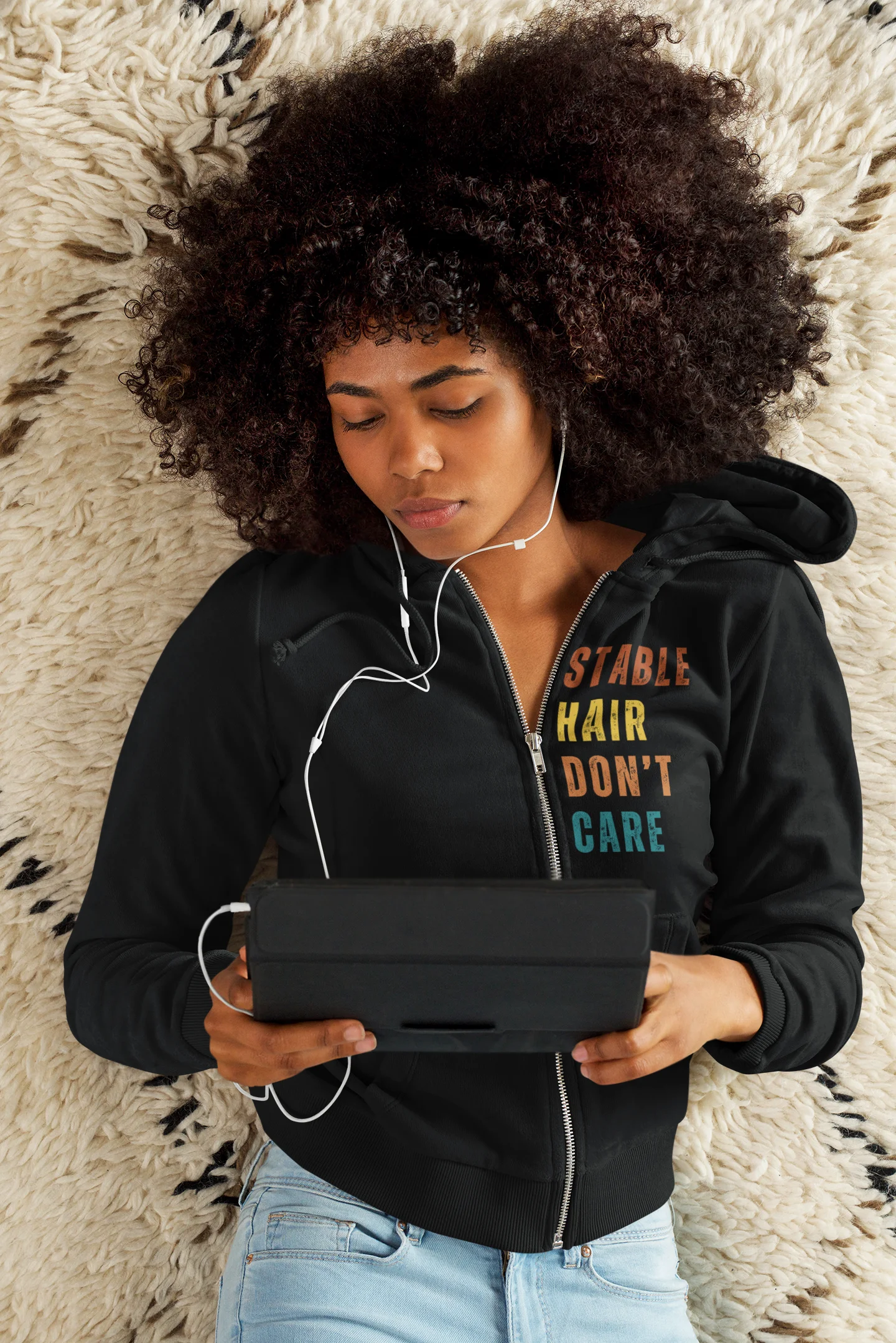 Equestrian Hoodie with Stable Hair Don't Care text and horse barn back print. Person wearing a black hoodie with colorful text, holding a tablet on a textured surface