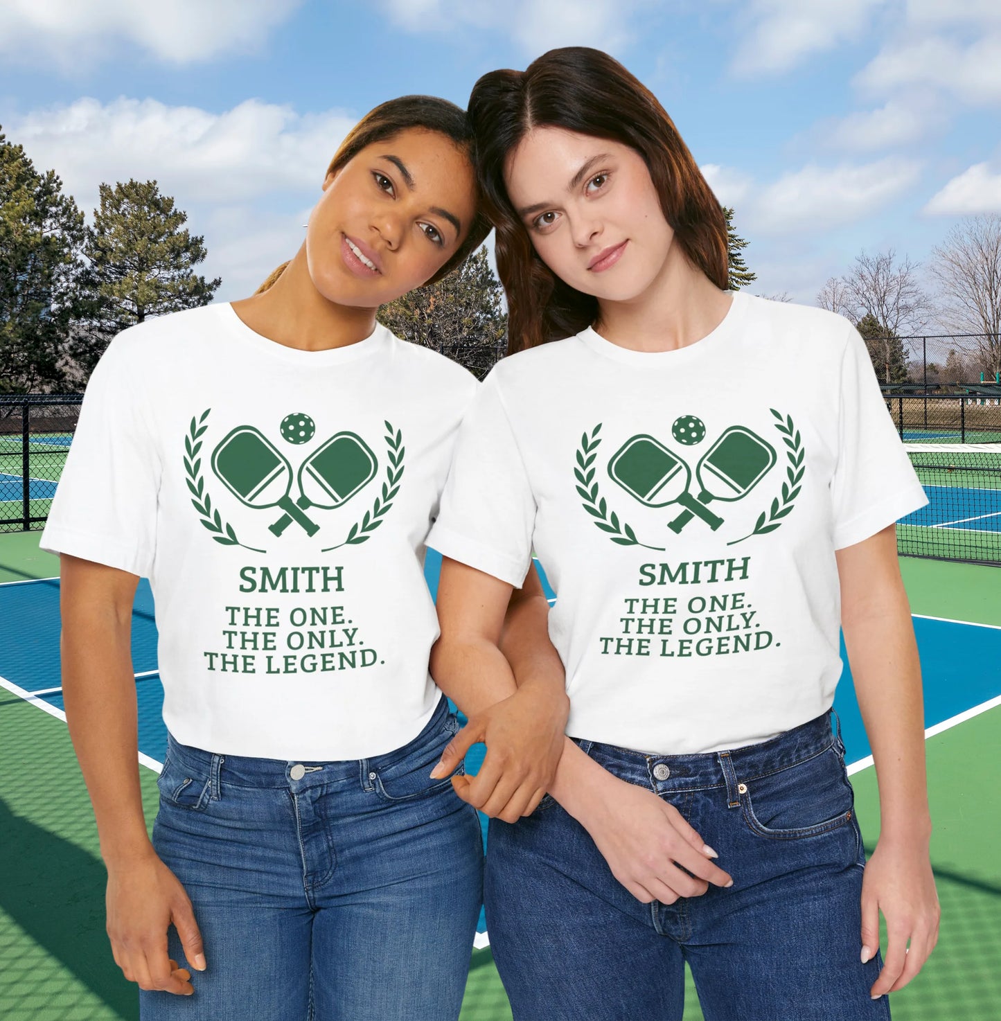 Funny pickleball shirt Two women standing together on a tennis court wearing matching white graphic t-shirts with a green crossed-racquet design and the text 'SMITH THE ONE. THE ONLY.