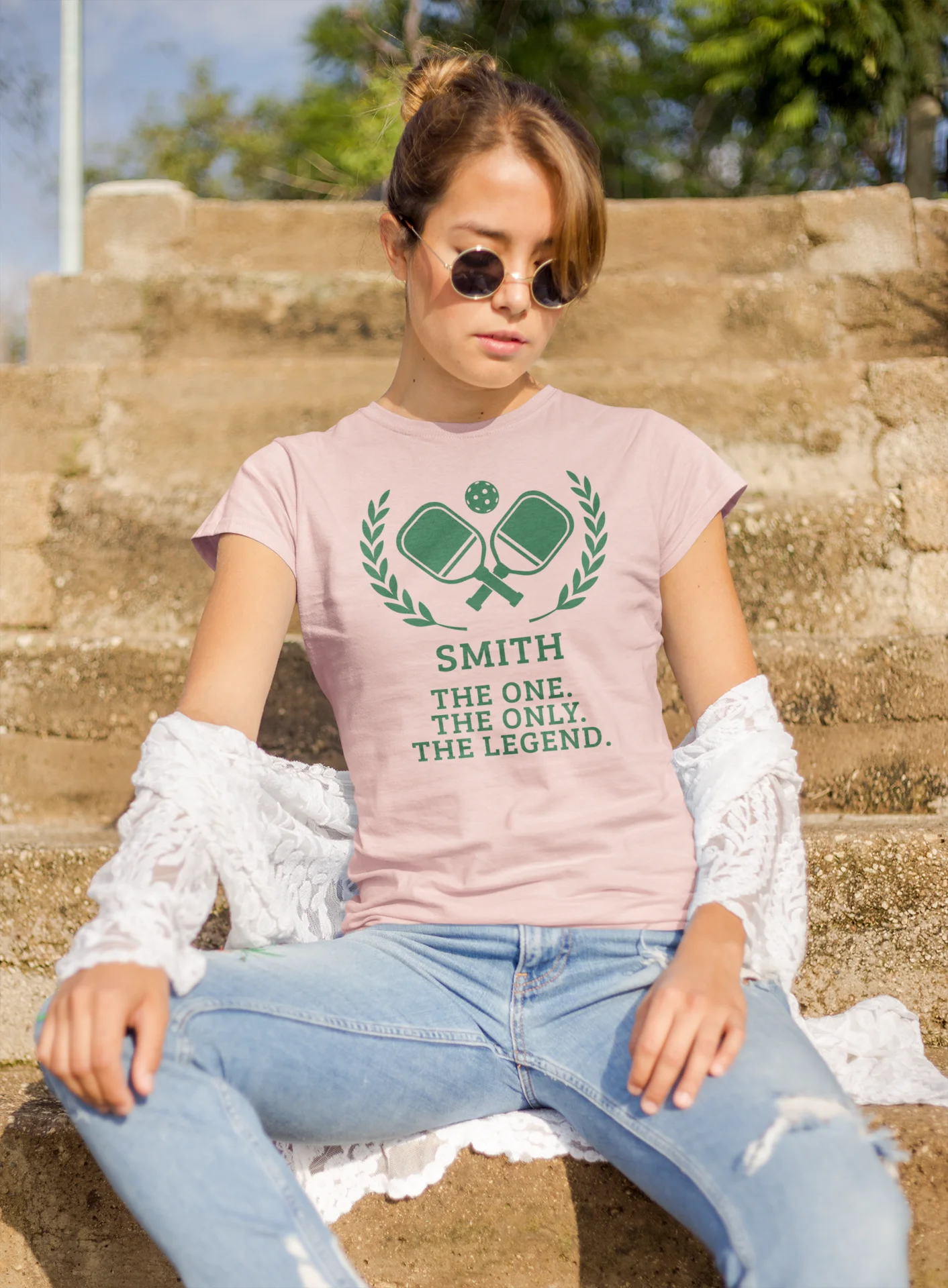 Funny pickleball shirt A woman sitting on outdoor stone steps wearing a light pink graphic T-shirt with a green tennis racket design and the text 'SMITH THE ONE. THE ONLY.