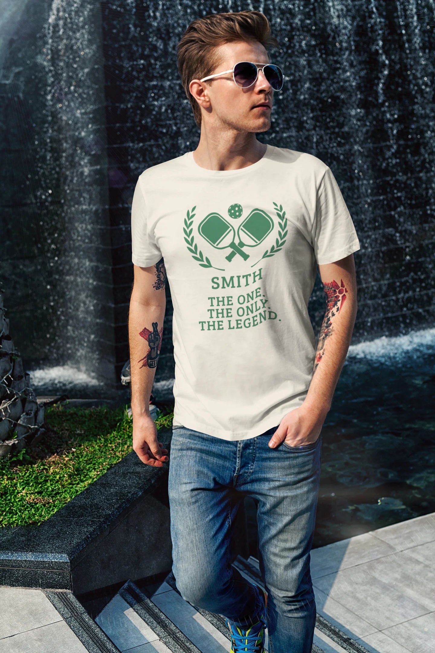 Funny pickleball shirt A man wearing a cream-colored t-shirt with a green graphic of crossed golf clubs and the text 'SMITH THE ONE, THE ONLY, THE LEGEND.' standing in front of a waterfall.