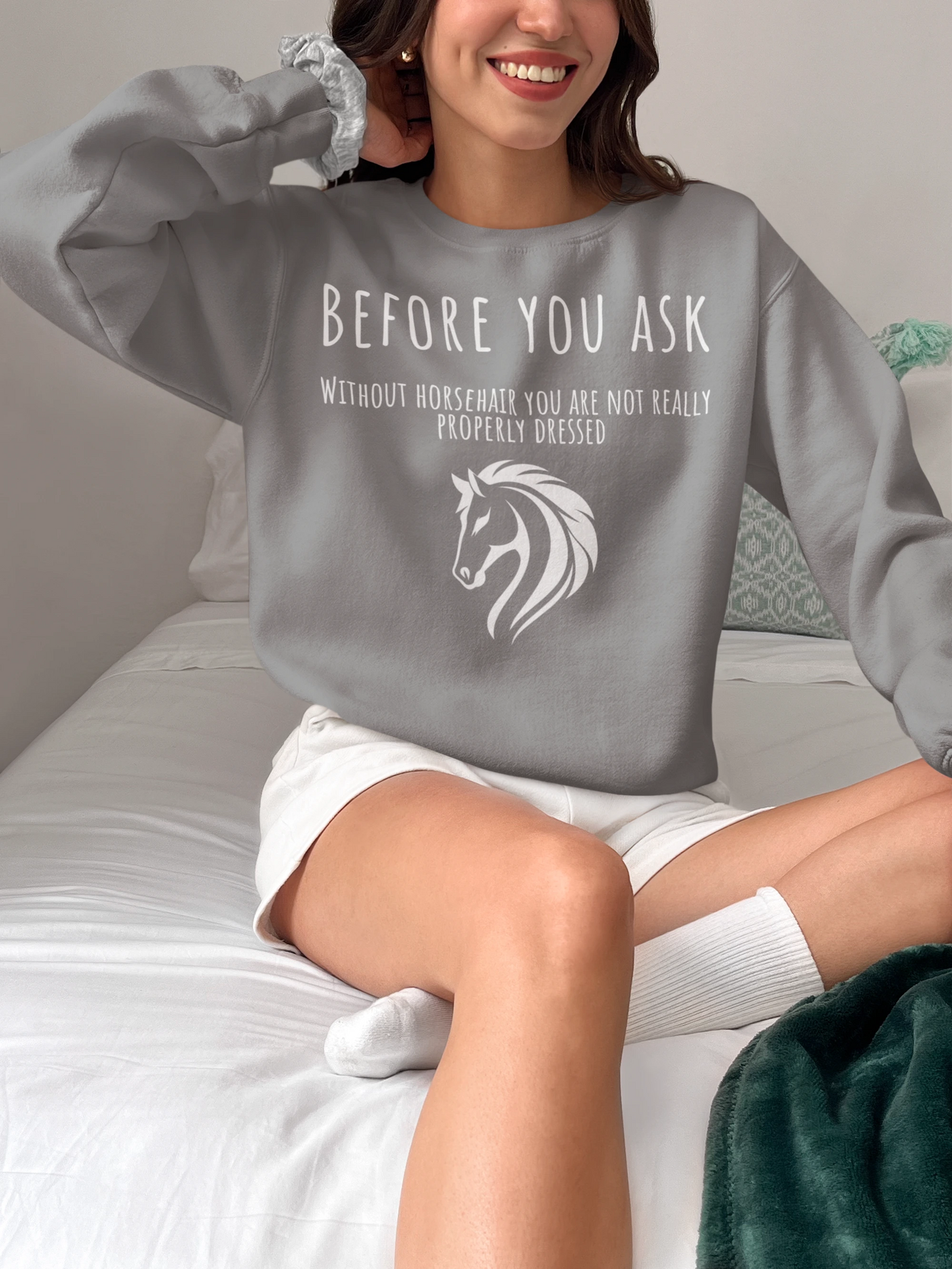 Person wearing a gray sweatshirt with a horse graphic and text, sitting on a bed.