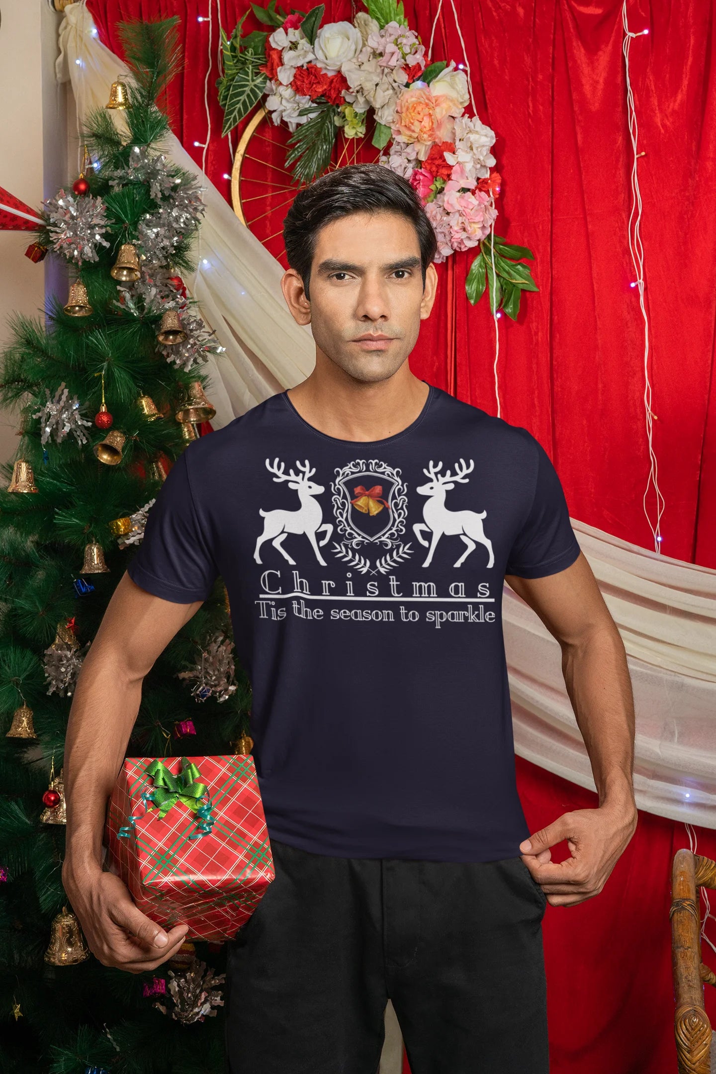 Man wearing a navy blue t-shirt with Christmas design in front of a decorated Christmas tree and red curtain.