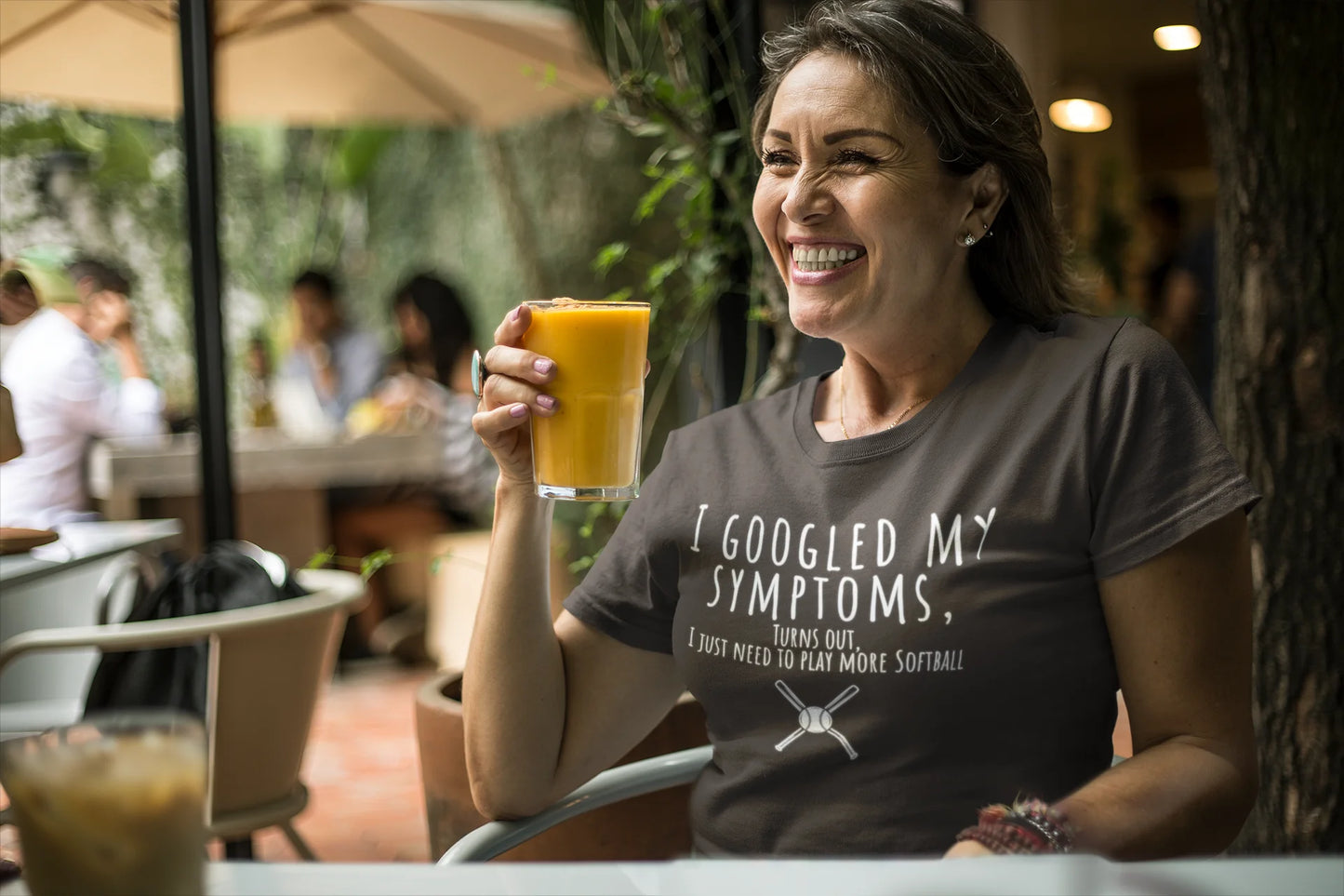 Woman sitting outdoors holding a glass of orange juice, wearing a t-shirt with humorous text.
