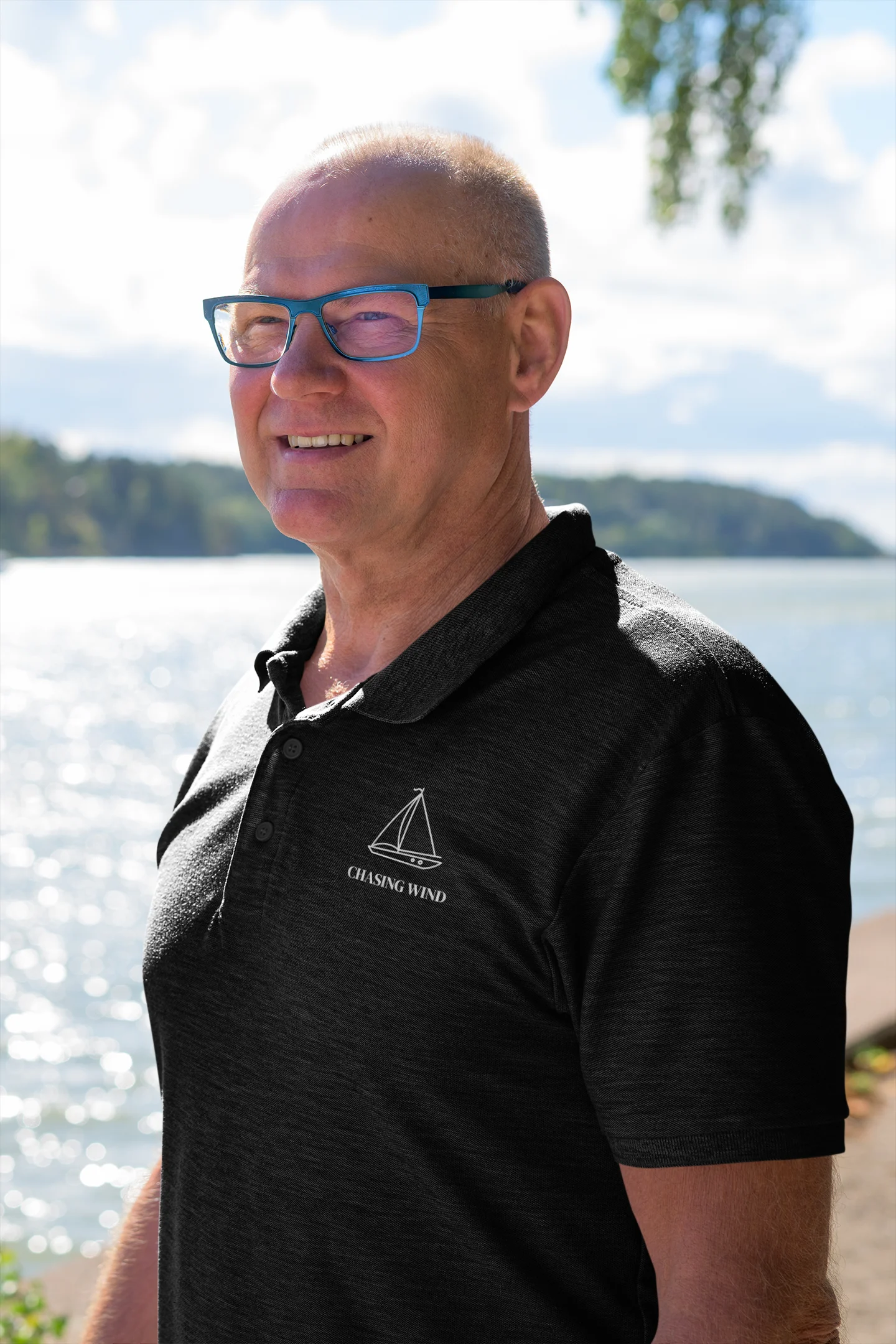 Sailing Polo Shirt Man wearing a black polo shirt with a logo standing by a body of water.