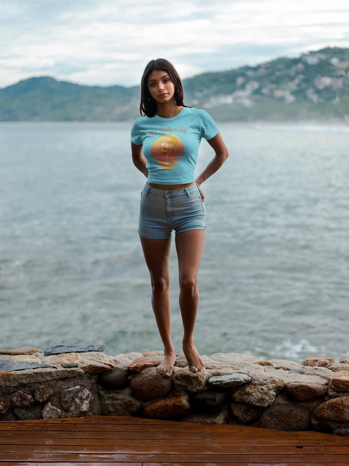 Aperol T-Shirt Woman wearing a light blue t-shirt with a graphic design, standing by a body of water with mountains in the background.